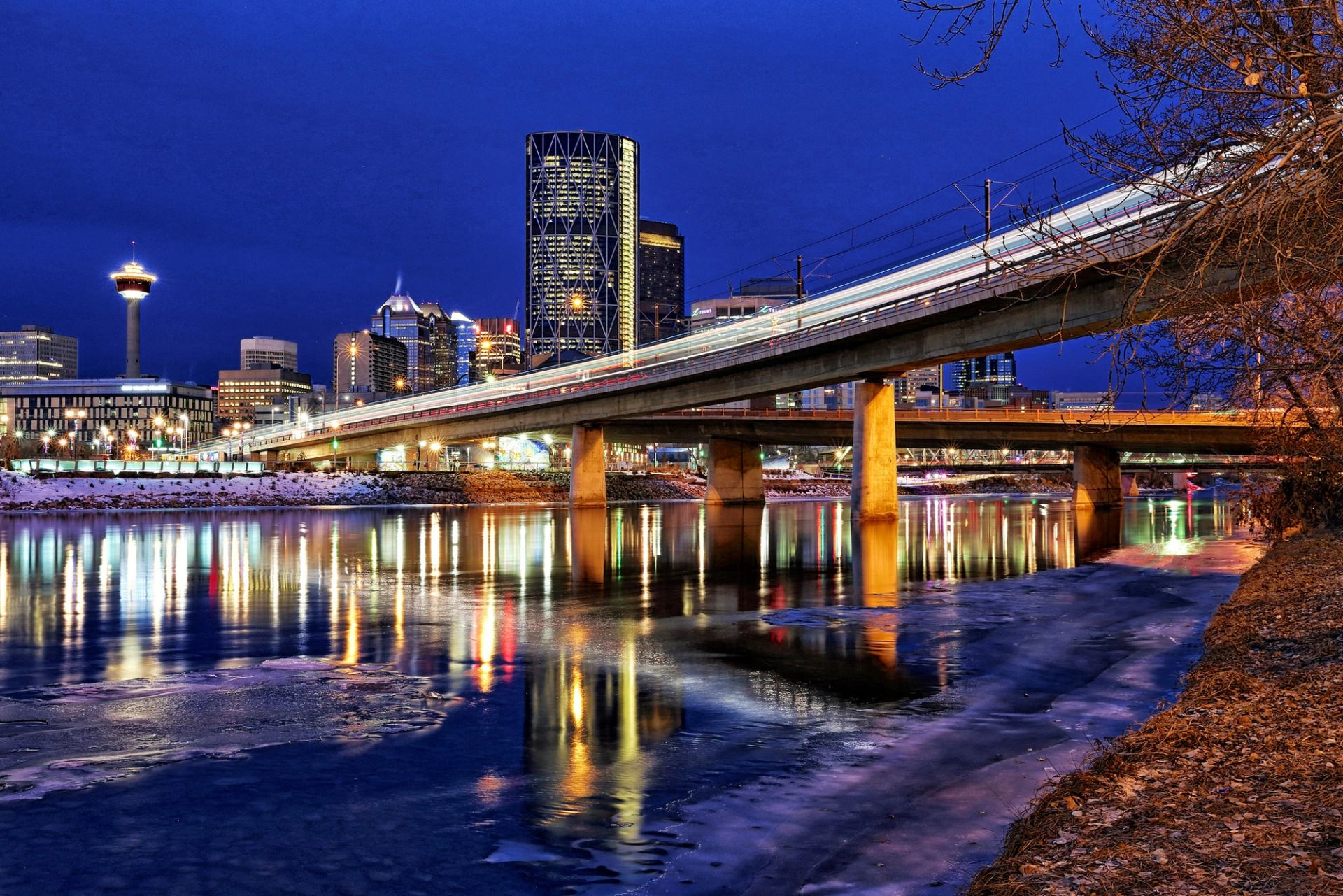 Lit-up bridge and city skyline reflected in water at night.