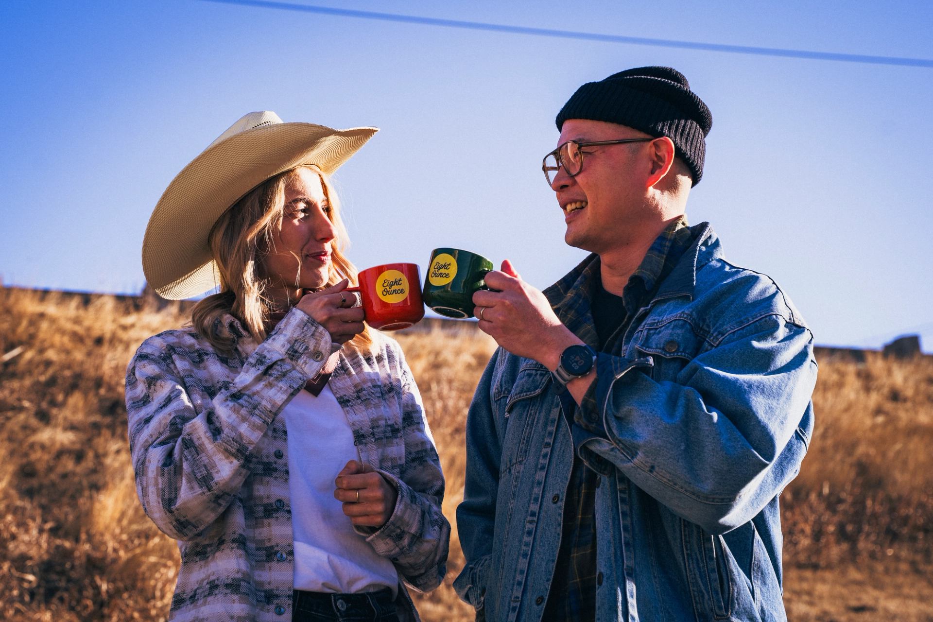 Two people outdoors clinking colorful coffee mugs in a western‑style setting with dry grass and blue sky.