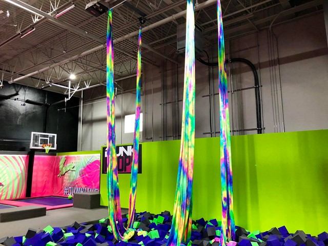 Colorful aerial silks hanging over a foam pit inside a trampoline park with bright neon walls.