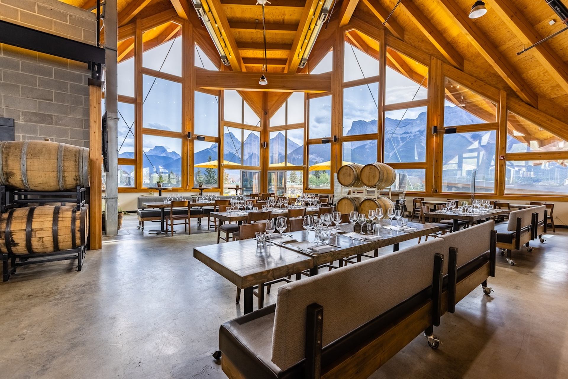 A restaurant with a vaulted wooden ceiling, large windows showcasing mountain views, set dining tables, and decorative wooden barrels.