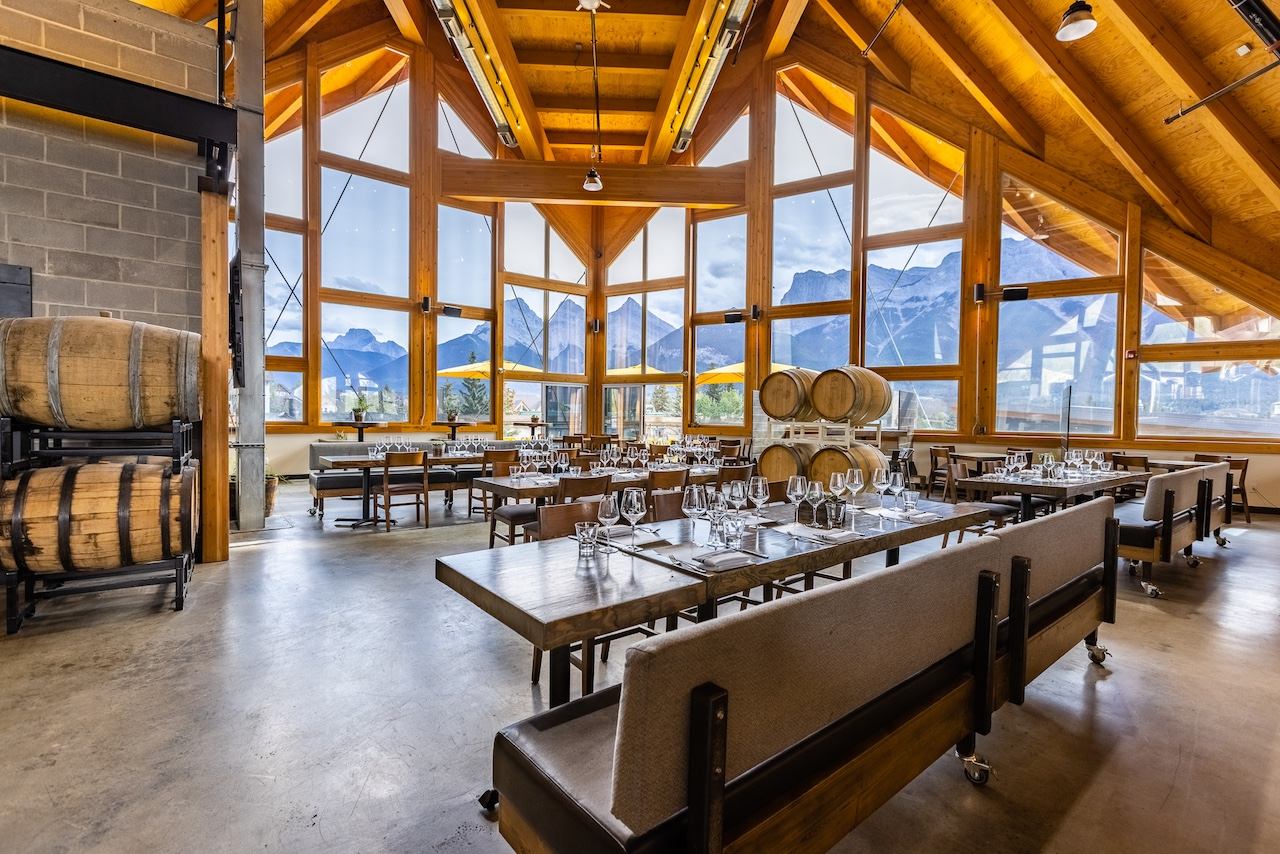 A restaurant with a vaulted wooden ceiling, large windows showcasing mountain views, set dining tables, and decorative wooden barrels.