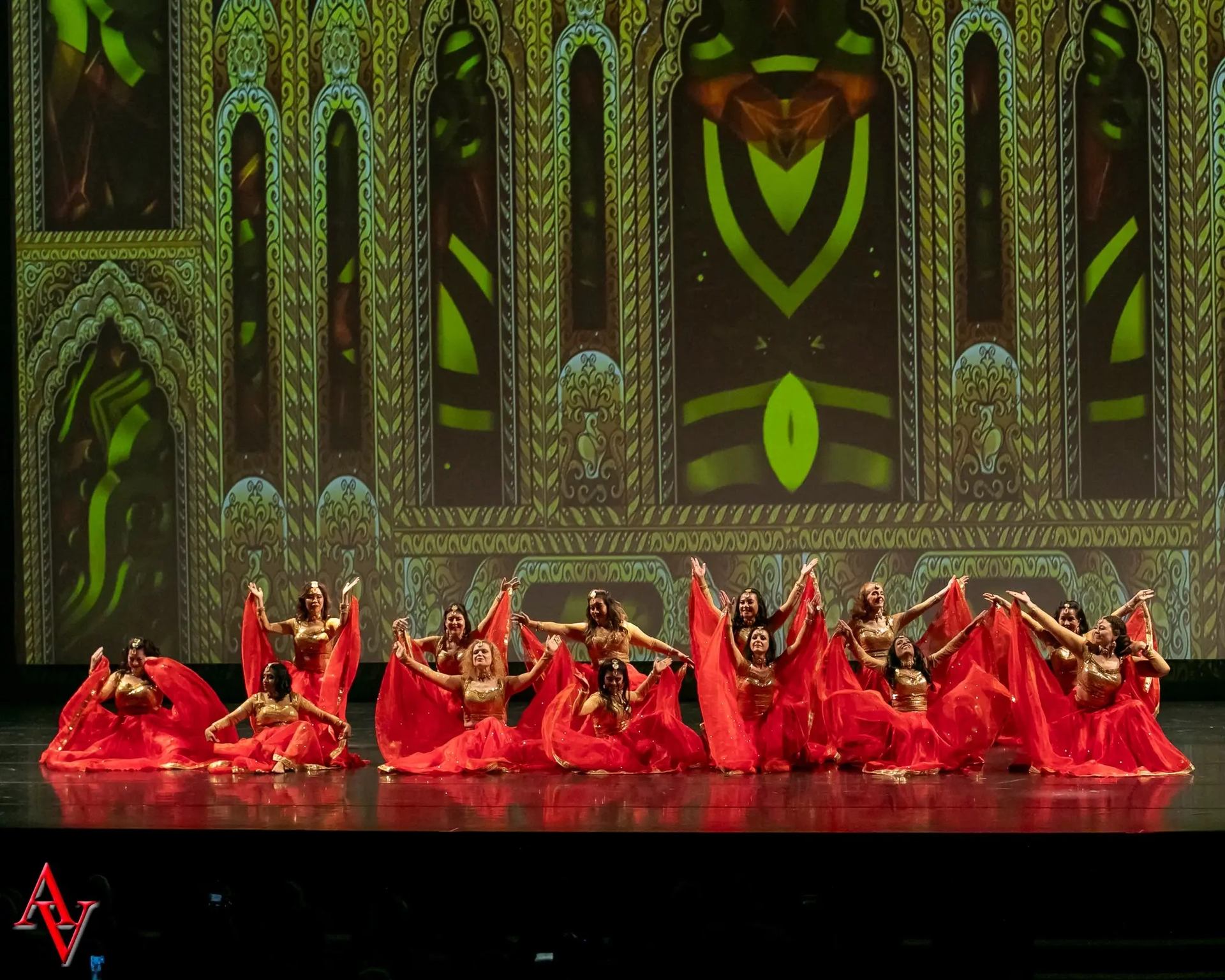 A group of dancers dressed in red and gold, seated and standing in a symmetrical formation, holding out sheer red fabric, with a grand, intricately patterned digital backdrop resembling palace architecture.