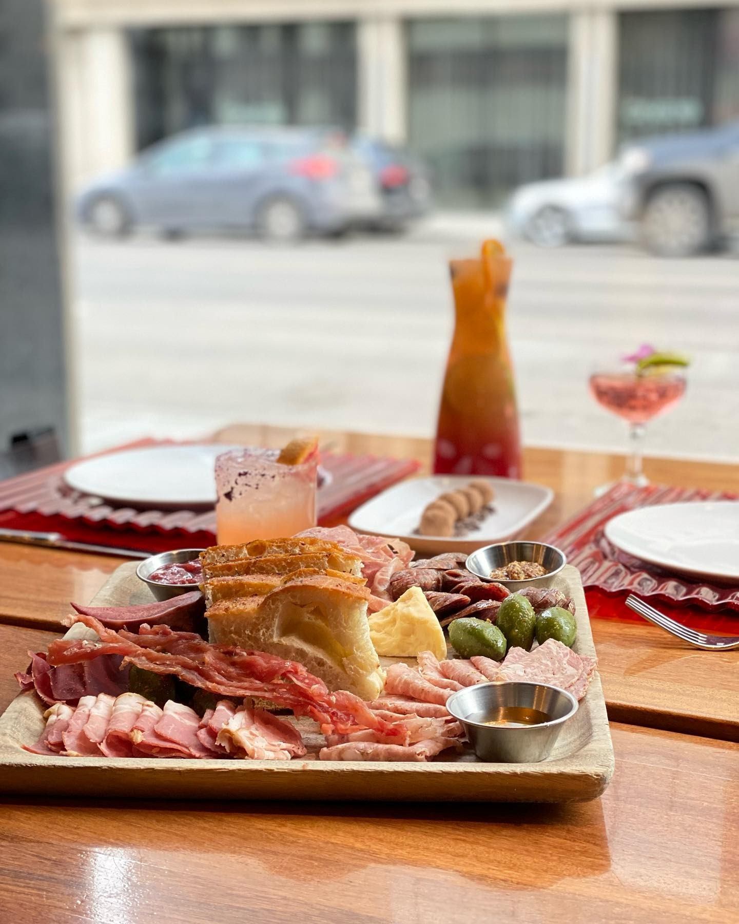 A vibrant charcuterie board with cured meats, cheeses, bread, and olives is on a wooden table alongside colorful cocktails, creating an inviting, social atmosphere.