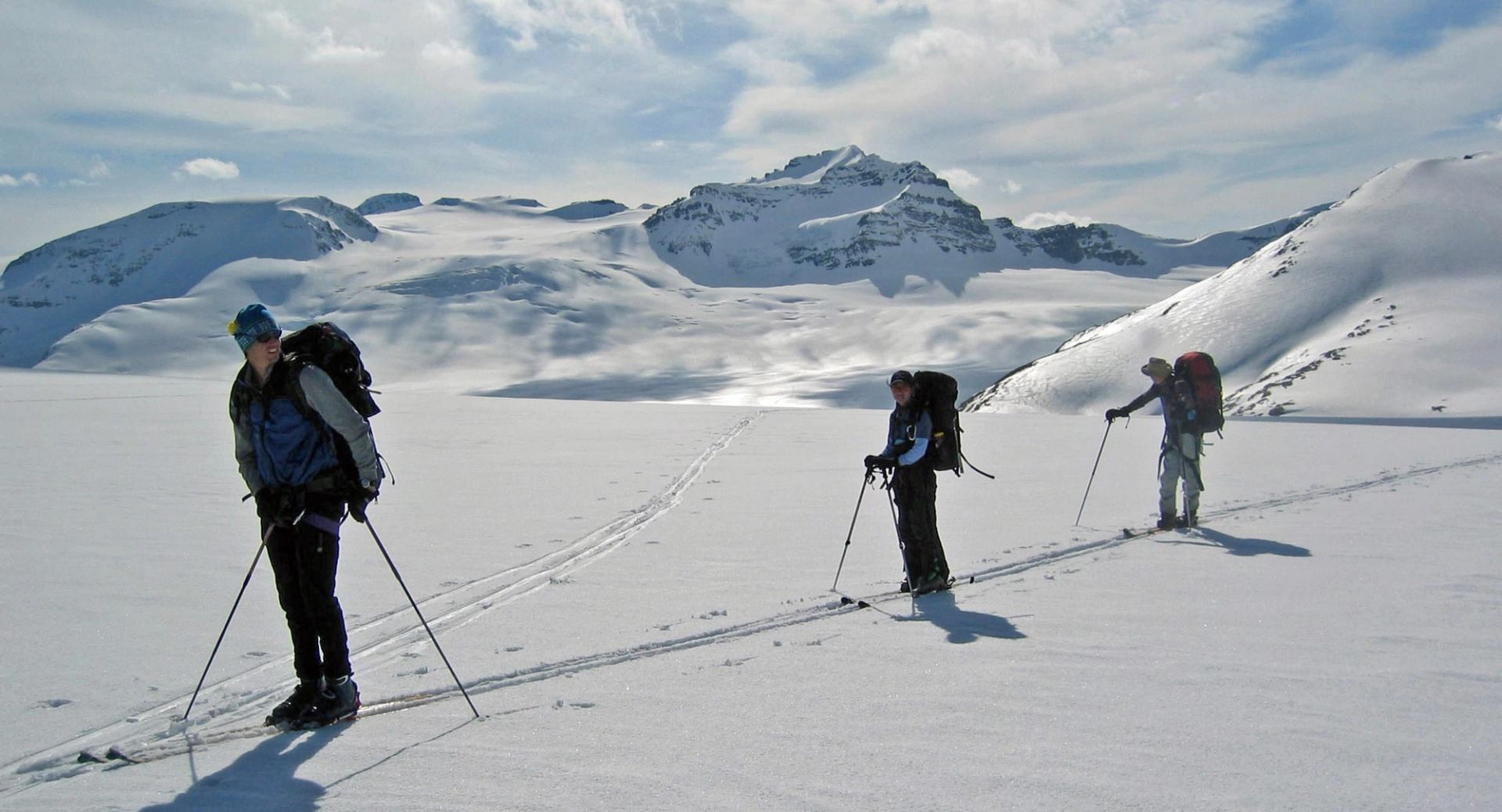 Three skiers cross a vast snowy expanse with packs and poles, surrounded by rugged peaks.