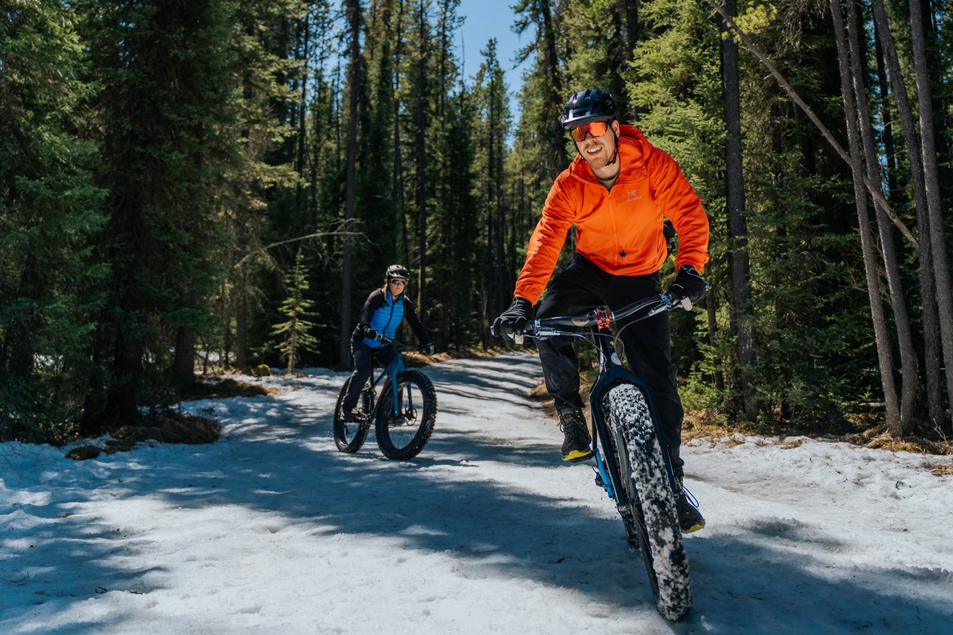 Mountain bikers ride a trail through a forest, one in orange jacket foreground.