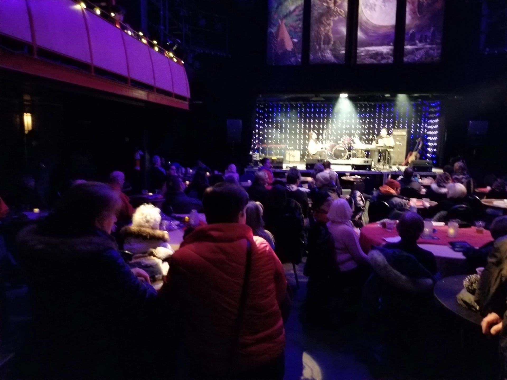 Performers on stage in cozy venue with audience seated at round tables.