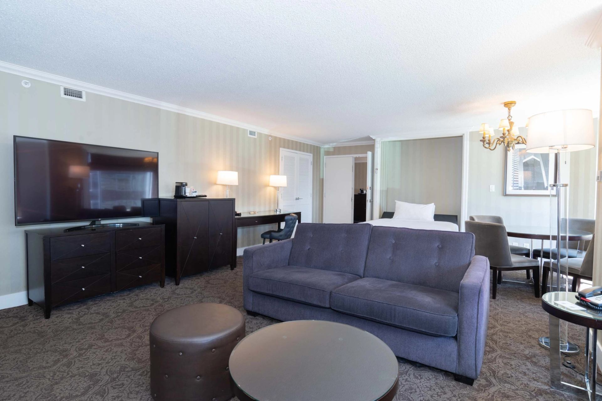 Hotel suite with grey sofa, TV, desk, bed, and dining table with chairs.