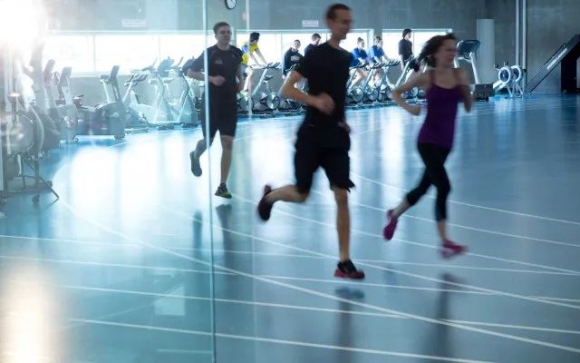 People run on an indoor track while cardio equipment lines the fitness area behind them.