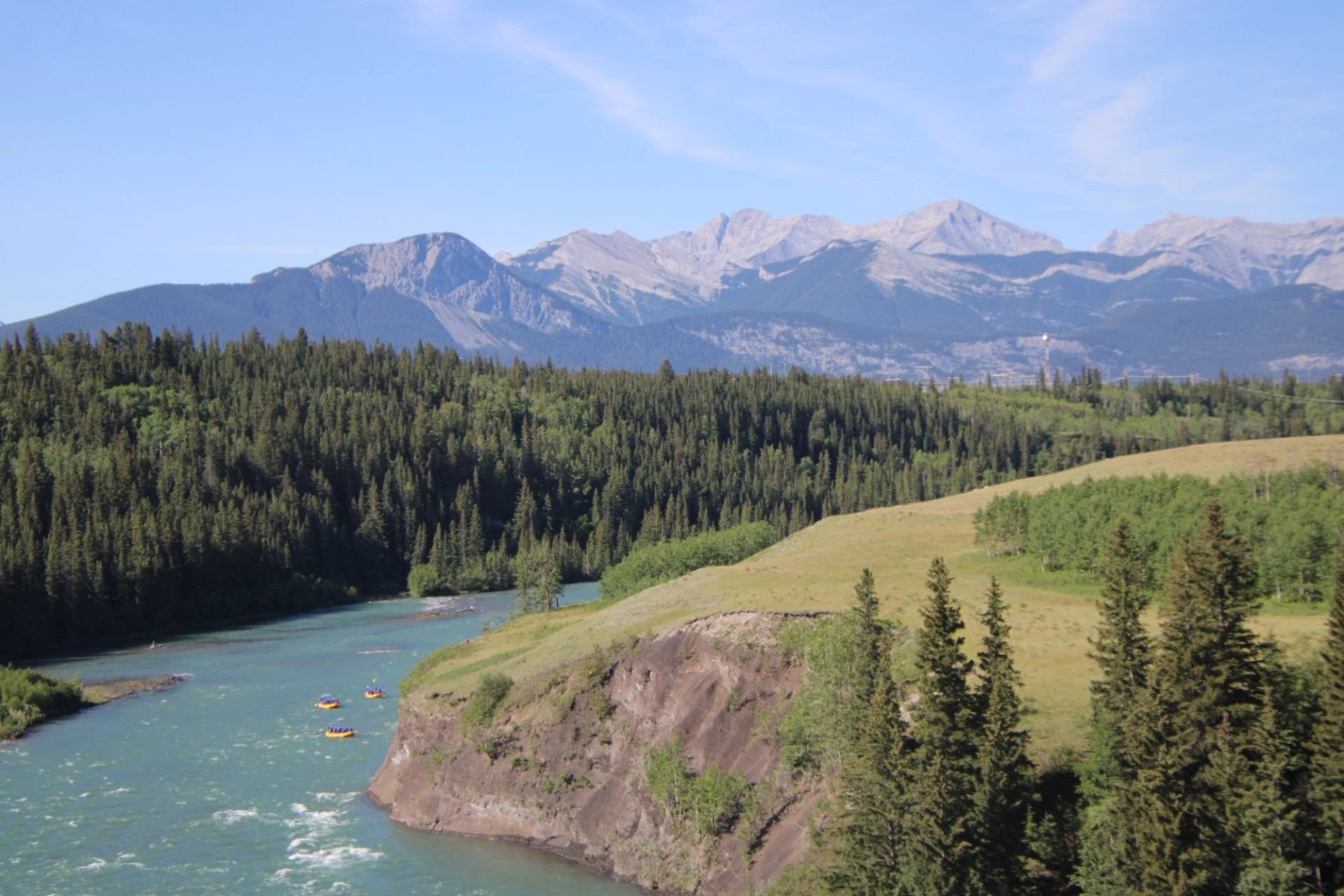 Mountain river with rafters flows past forested slopes and grassy cliffs under clear sky.