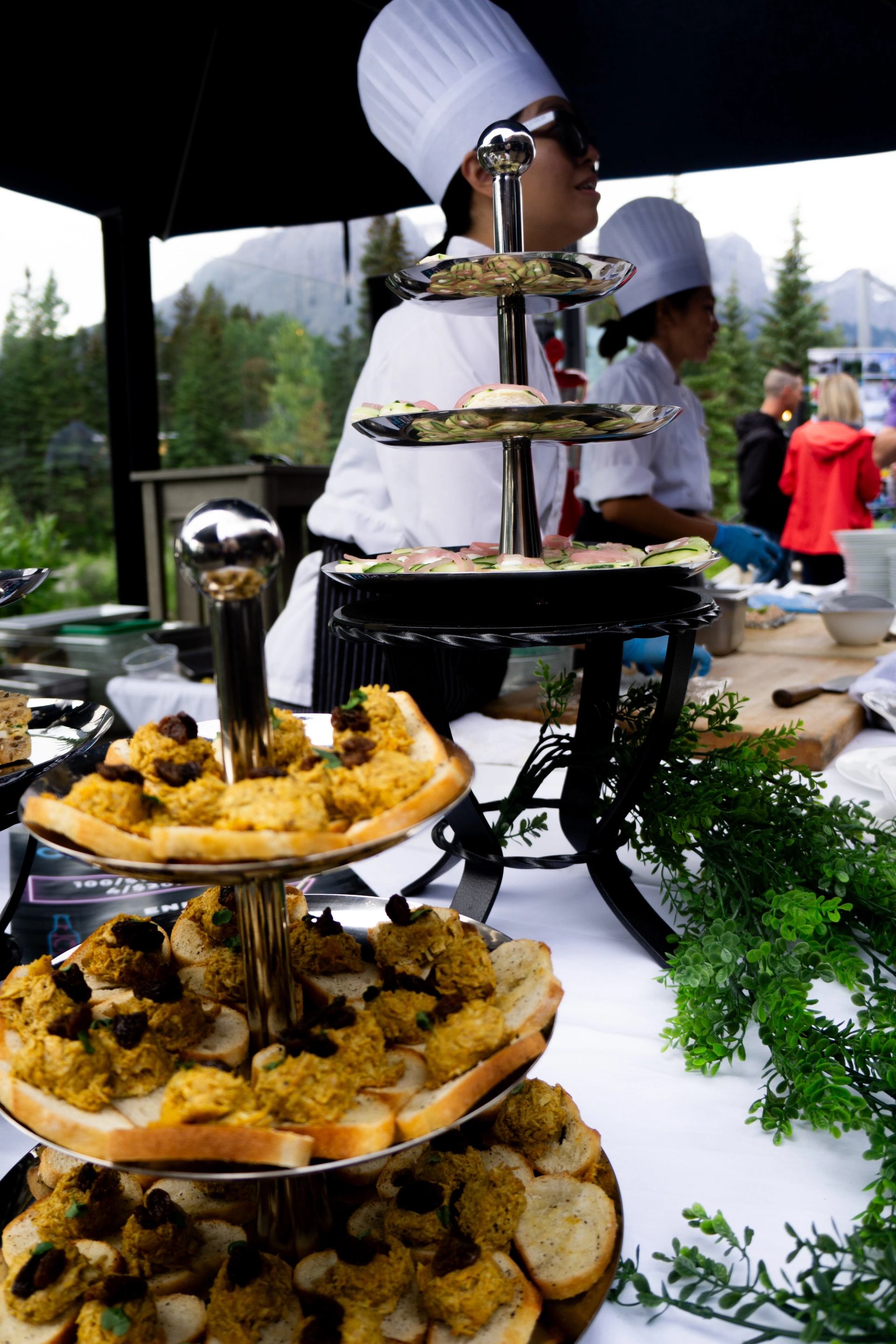 Tiered trays of appetizers on outdoor buffet table with chefs and guests in background.