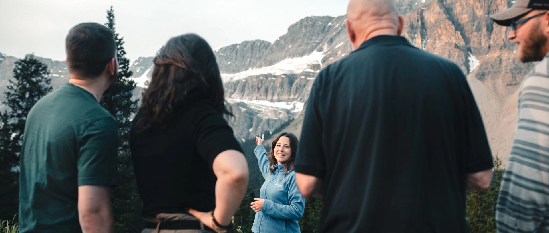 Guide in blue jacket points toward rocky, snow-patched mountain while group watches.