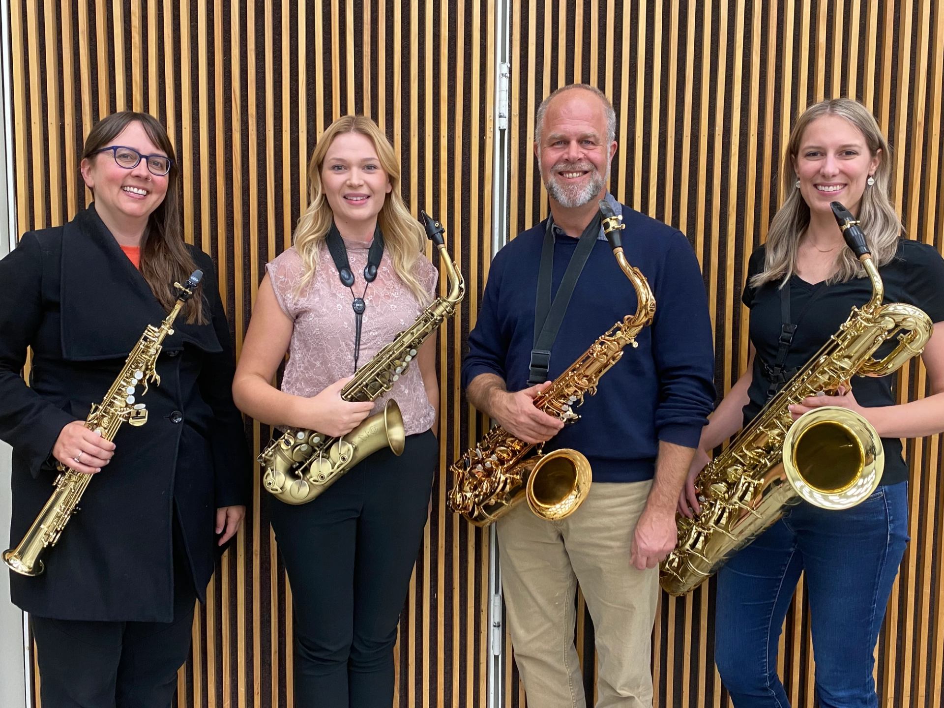 Four saxophone players stand side by side holding instruments against a wooden slat wall.