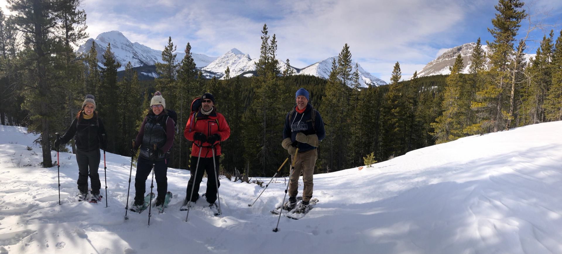 Four hikers on snowshoes standing on a snowy slope with forest and Rocky Mountains behind.