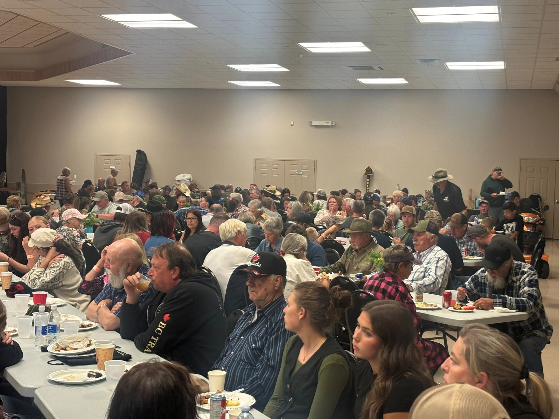 Crowded community hall with people seated at long tables eating and talking during event.