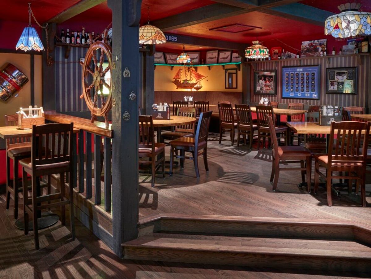 A dining area with wooden tables and chairs features nautical décor, including a ship’s wheel and model sailboat on the wall. Warm lighting and wood accents create a casual, themed restaurant setting.