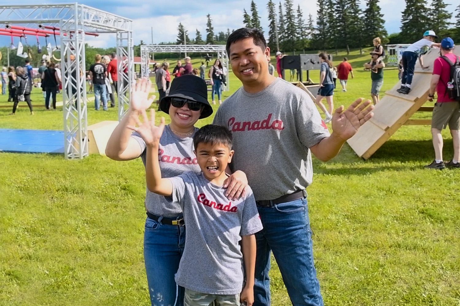 Three people in “Canada” shirts wave on a grassy field at an outdoor event in Wainwright.