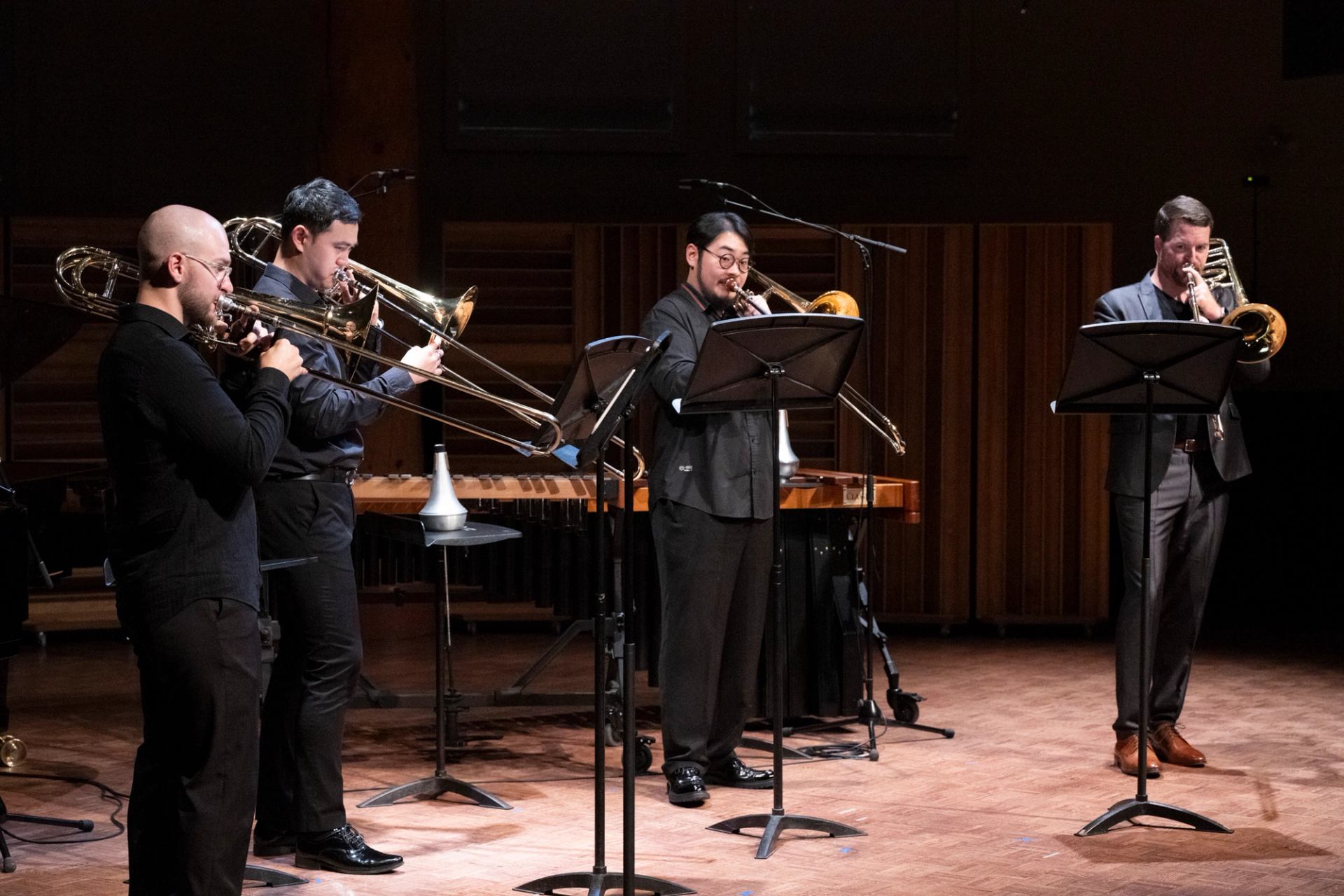 Four trombone players perform onstage with music stands and percussion instruments behind them.