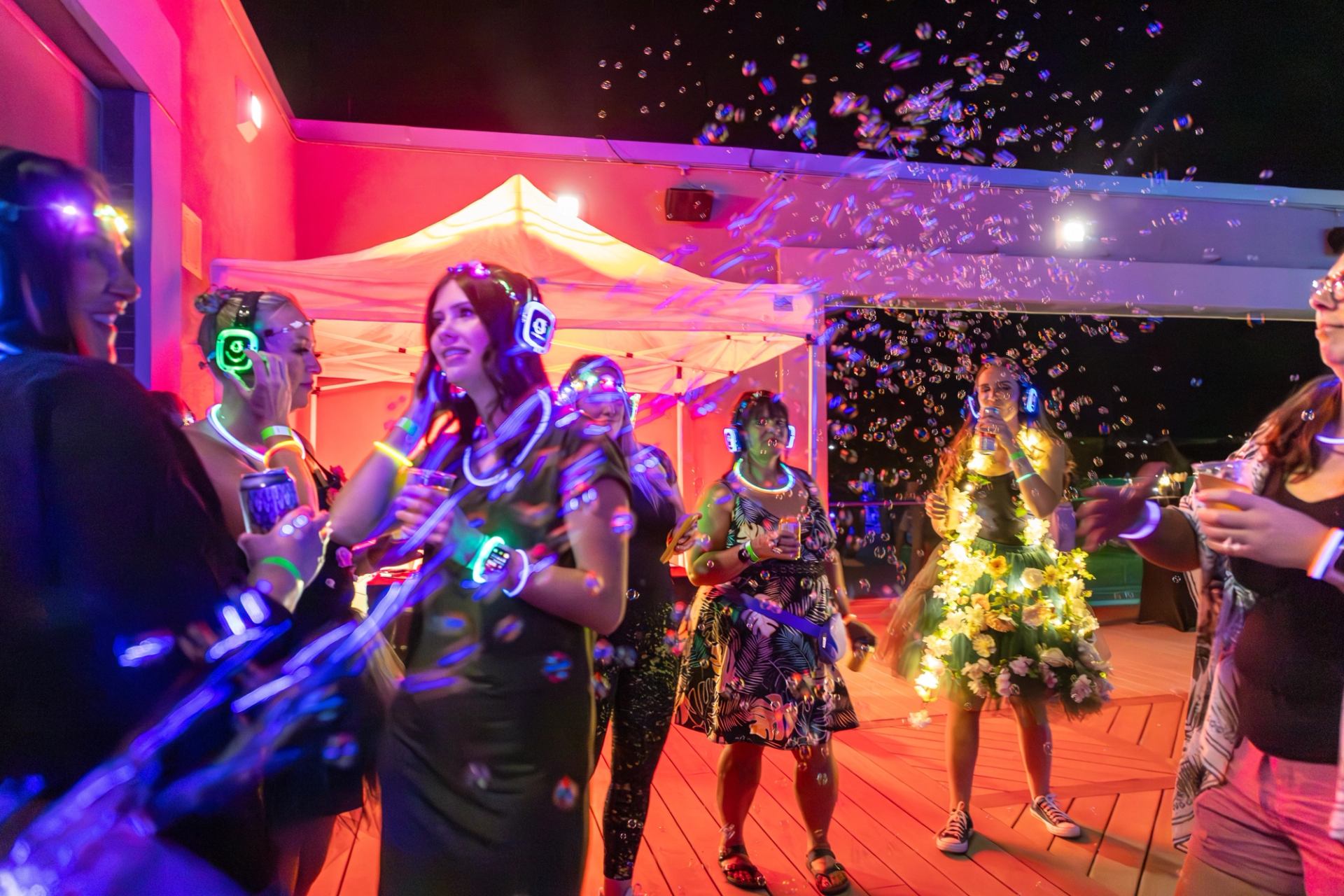 People wearing glowing headphones and accessories dance among bubbles under colourful lights.