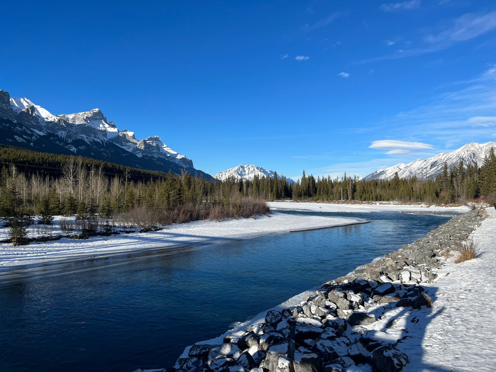 Snow-covered riverbank with evergreen forest and distant rocky mountains under clear blue sky.