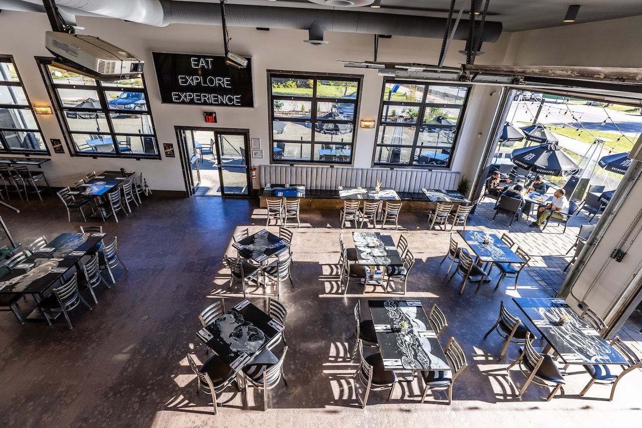 An aerial view of a modern restaurant interior with many empty tables, large windows, an open garage door leading to an outdoor patio with seated guests and umbrellas, and a sign reading "EAT EXPLORE EXPERIENCE."