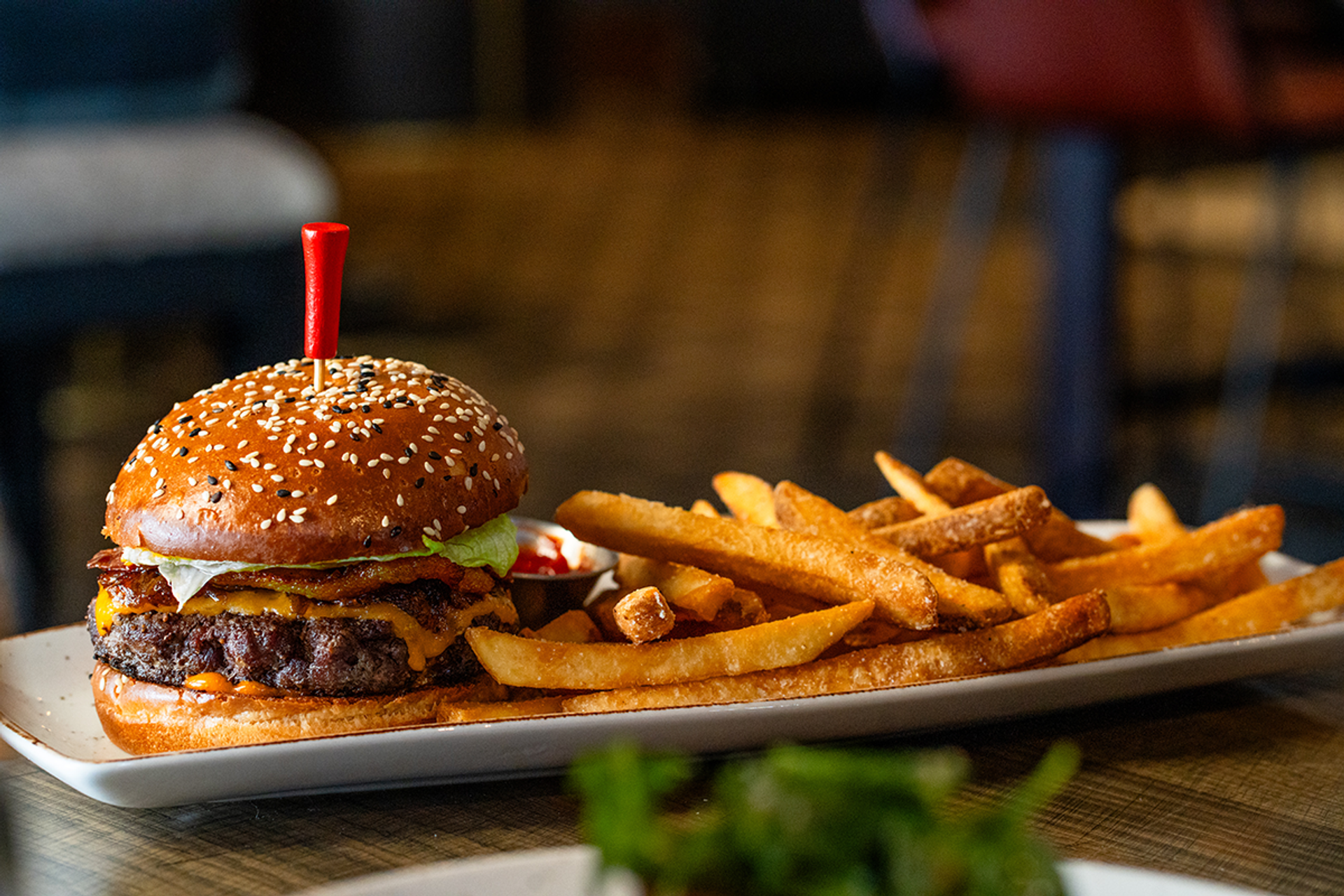 Burger with sesame bun and fries on rectangular plate in restaurant setting.