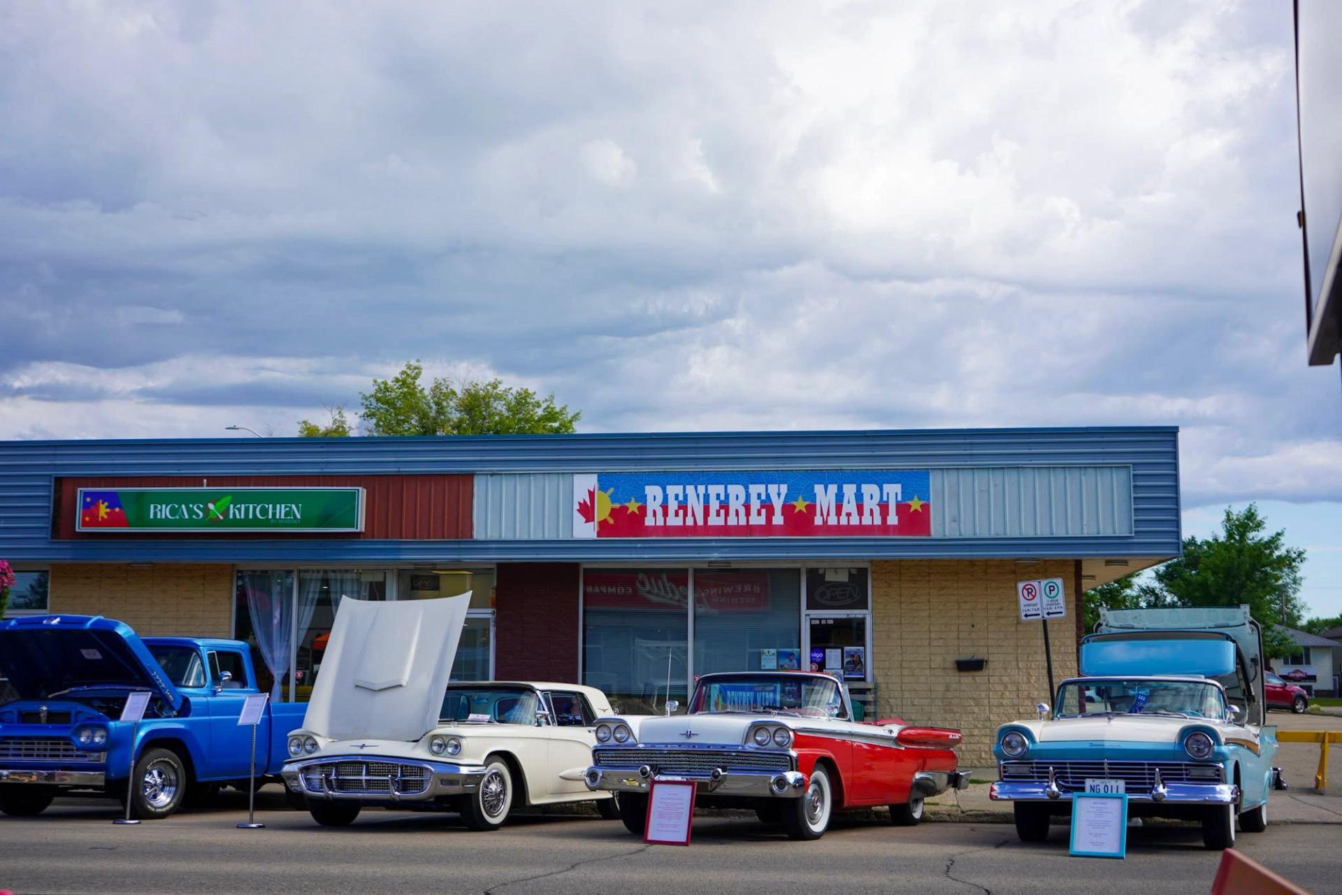 Classic cars with open hoods parked outside Renerey Mart and Rica’s Kitchen storefronts.
