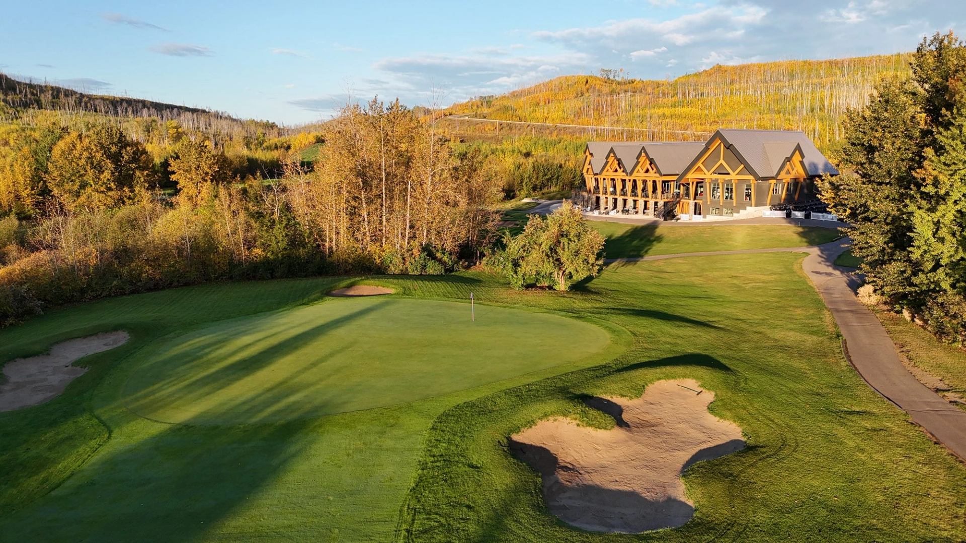 A green with bunkers near the large wooden clubhouse surrounded by fall foliage.