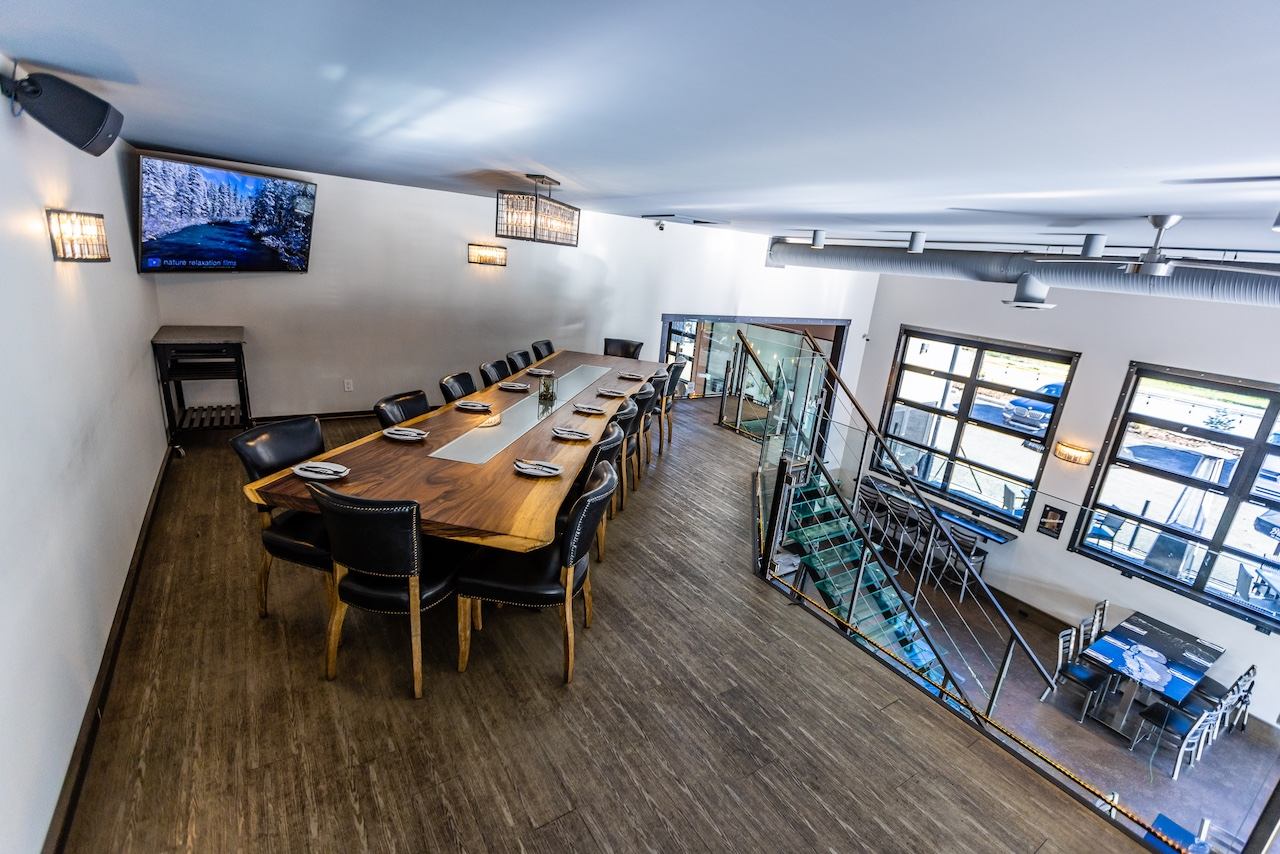 A modern private dining room with a long wooden table, black chairs, a wall-mounted TV, and a glass staircase leading to a lower level with large windows.