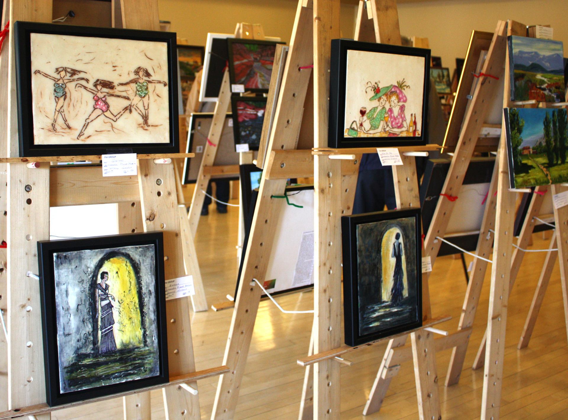 Wooden easels display framed paintings in a brightly lit indoor art exhibition.