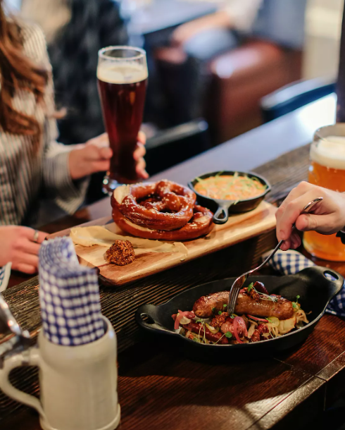 Pretzel, sausage entrée, and glasses of beer are served on a wooden table inside the restaurant.