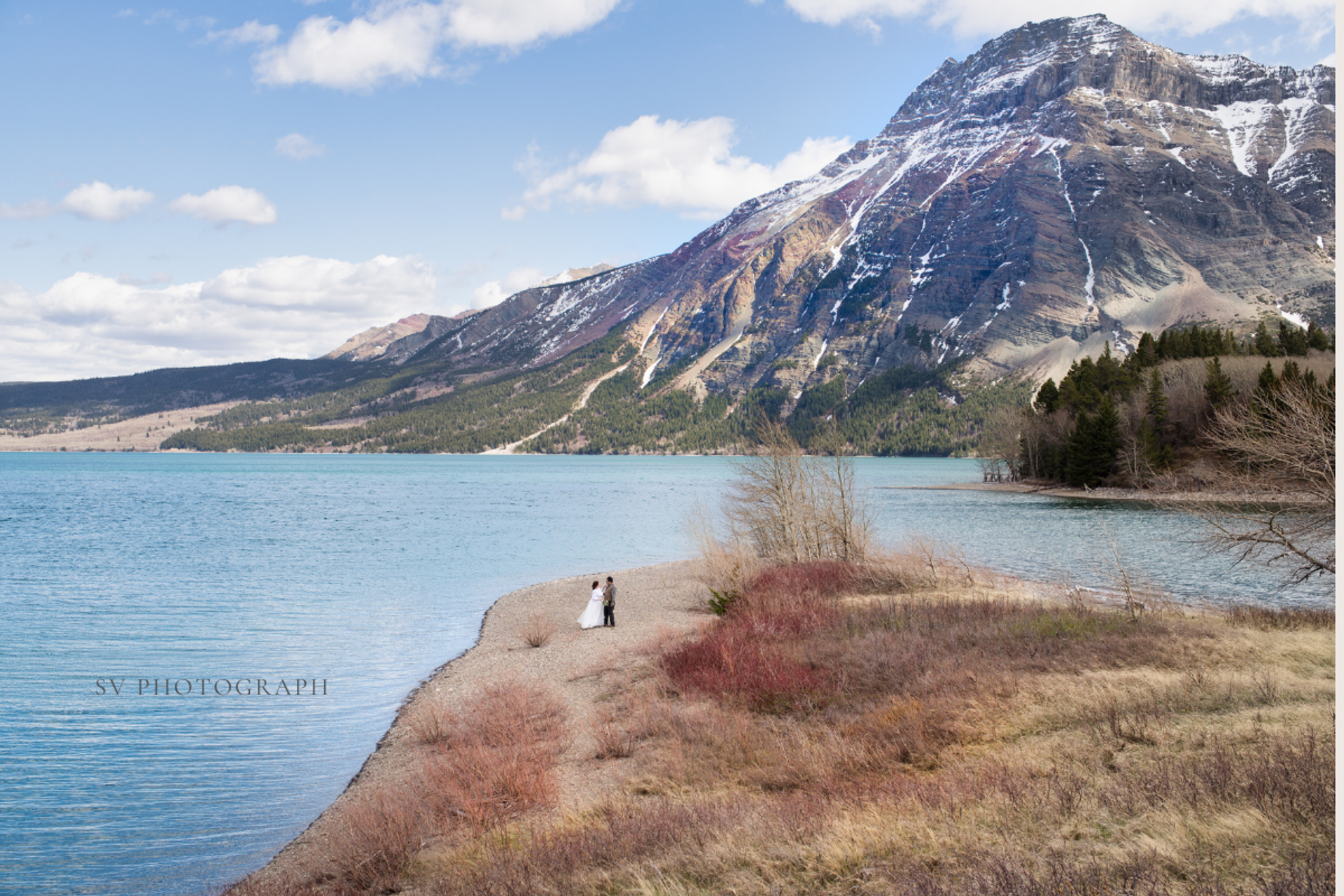Couple standing on a narrow lakeshore with blue water and a snow-dusted mountain behind.