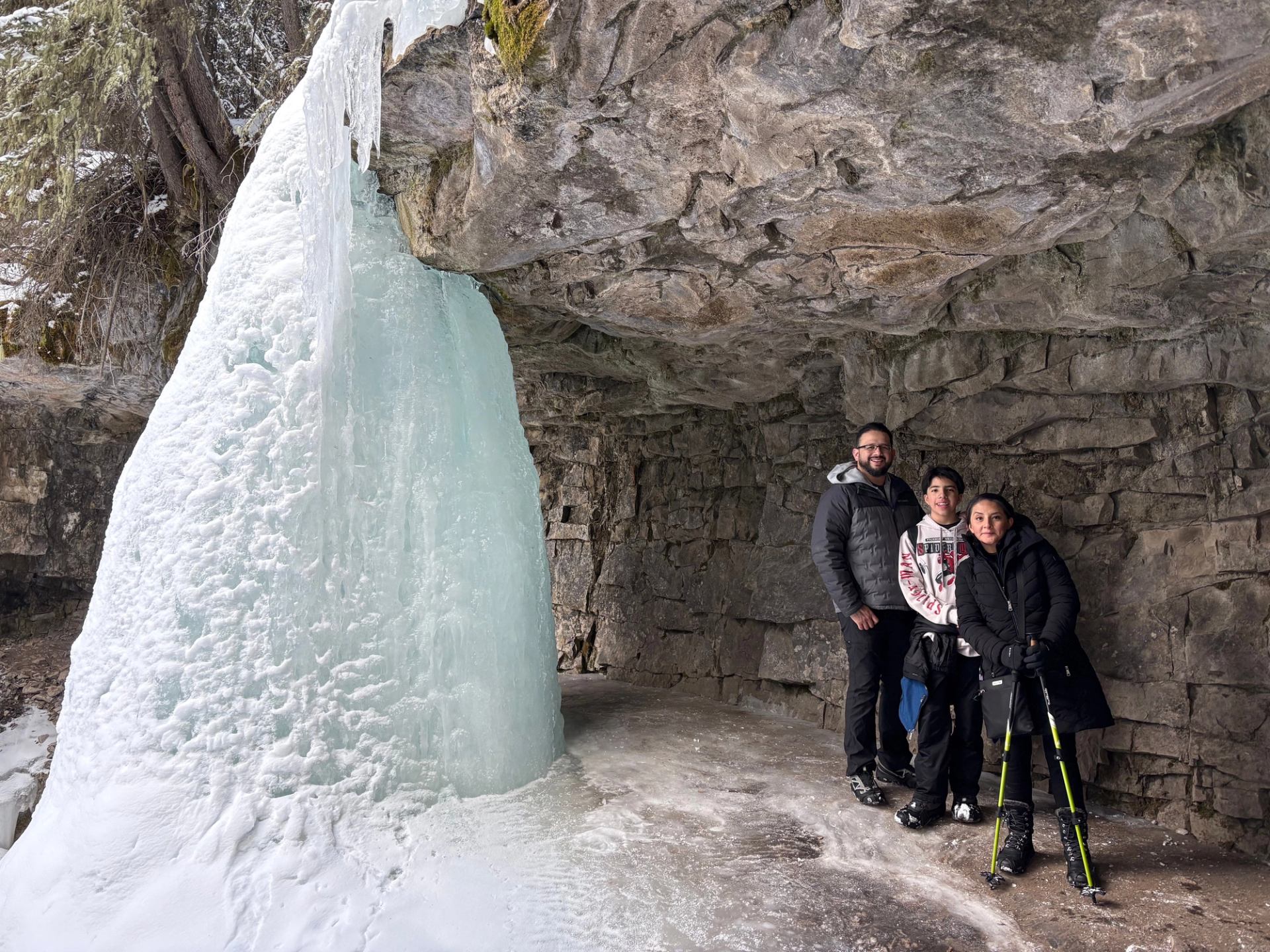 Large frozen waterfall beside rocky overhang with three bundled hikers standing on ice.