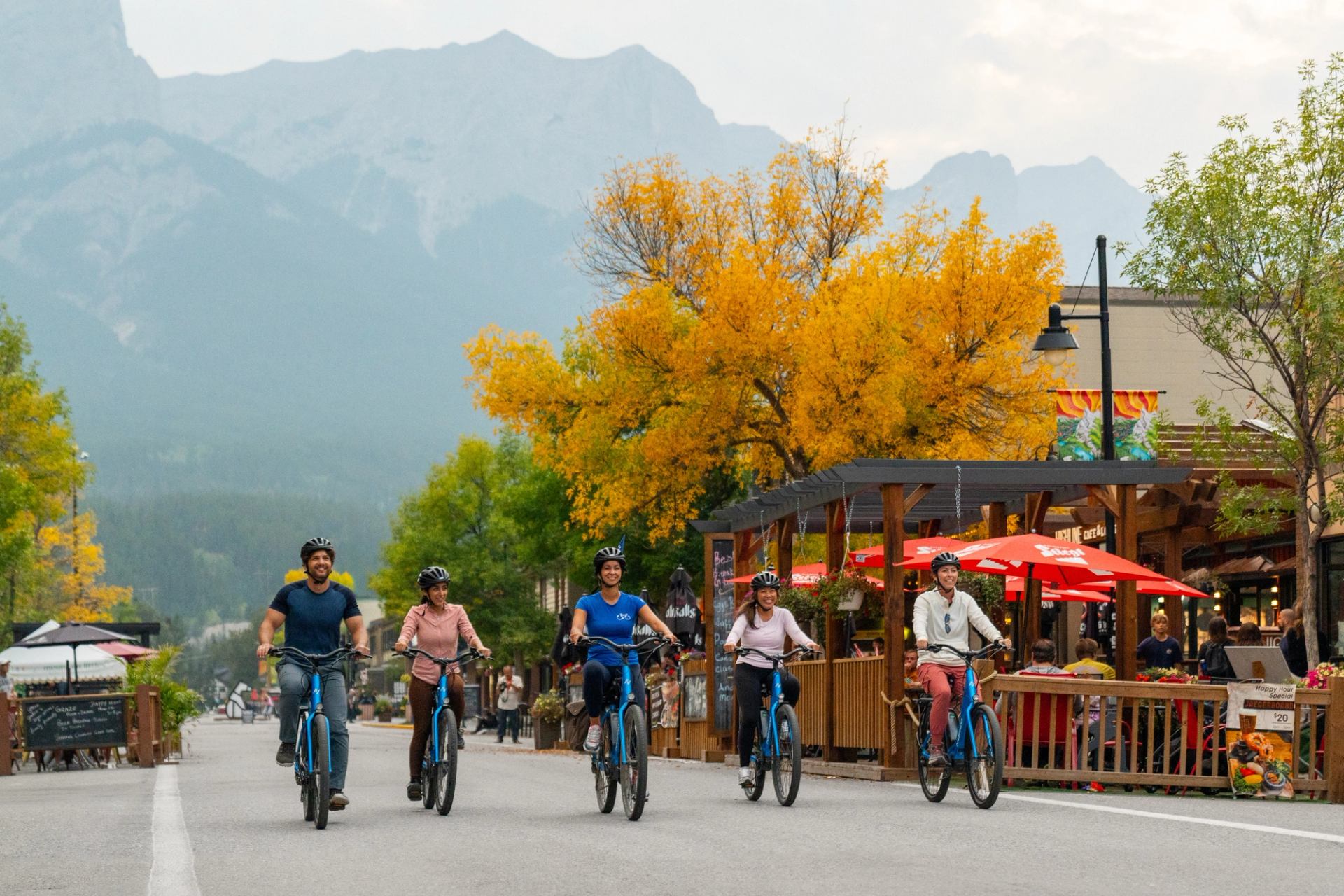 Five people riding bikes on a street lined with autumn trees and outdoor cafes, with mountains in the background.