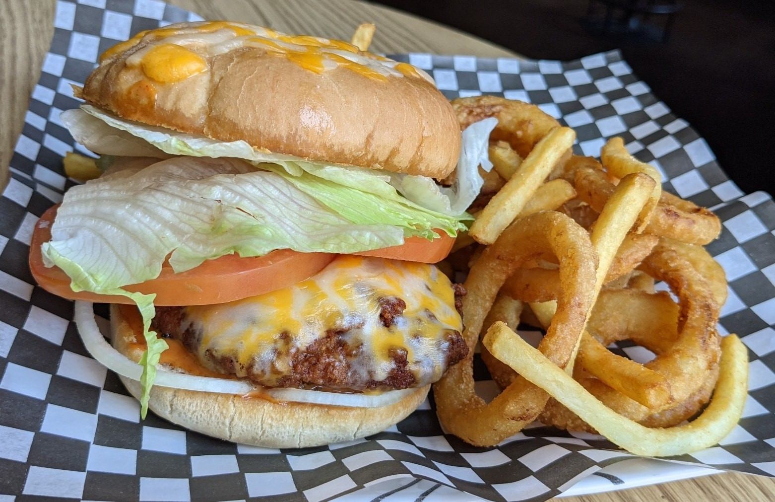 Cheeseburger with lettuce and tomato beside onion rings and fries on checkered paper.