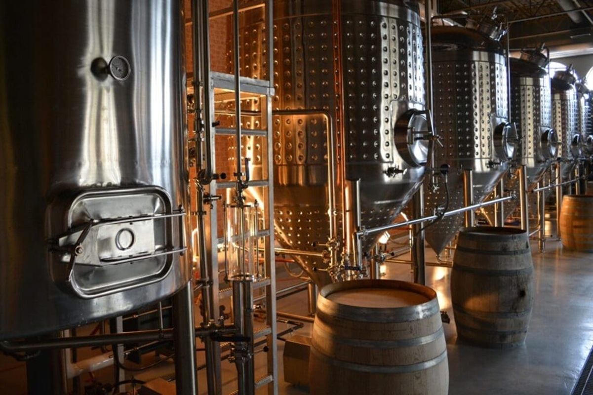 Stainless steel fermentation tanks and wooden barrels inside an industrial brewery facility.