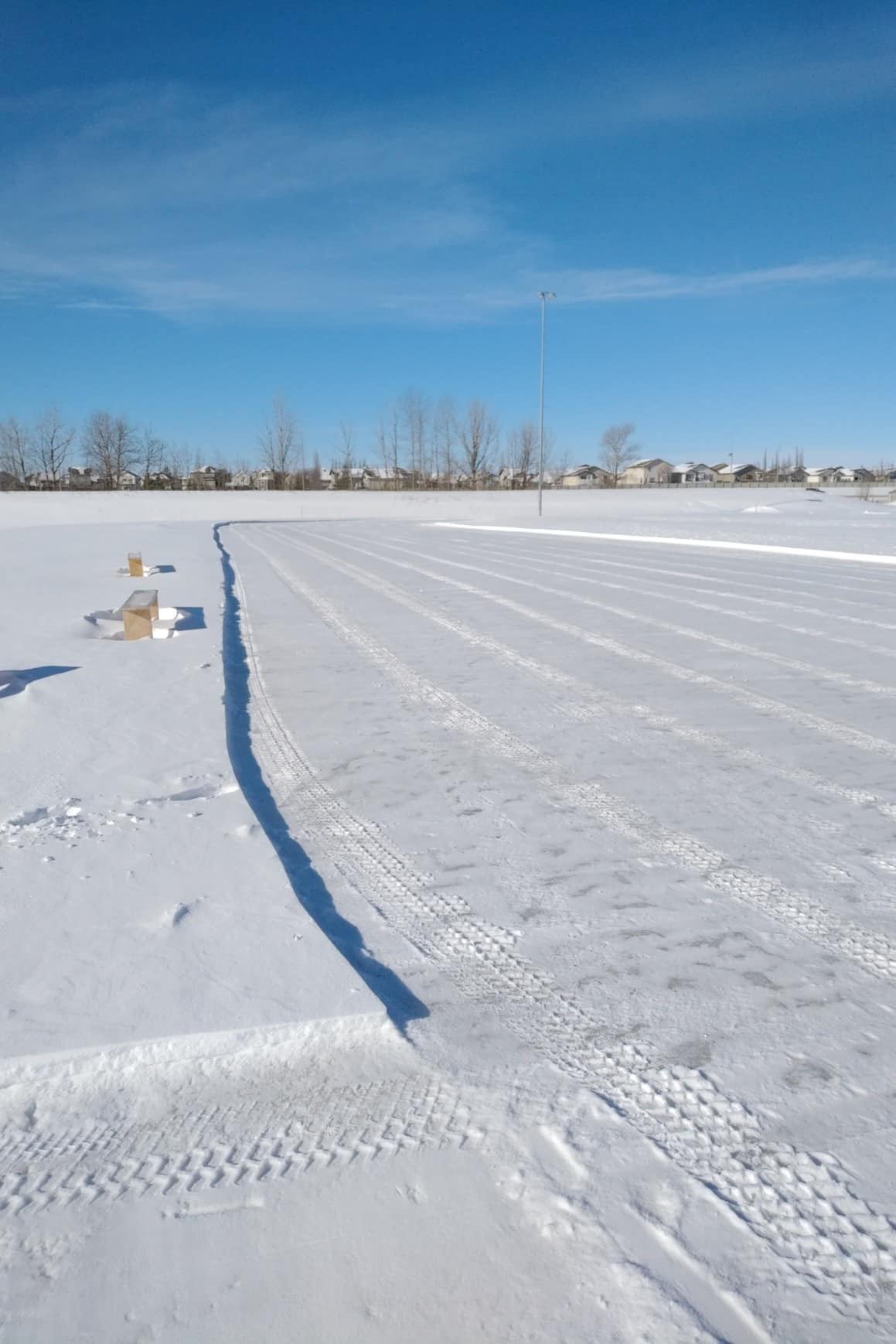 Skating oval on a snowy outdoor rink with marked tire tracks and boards along edges.