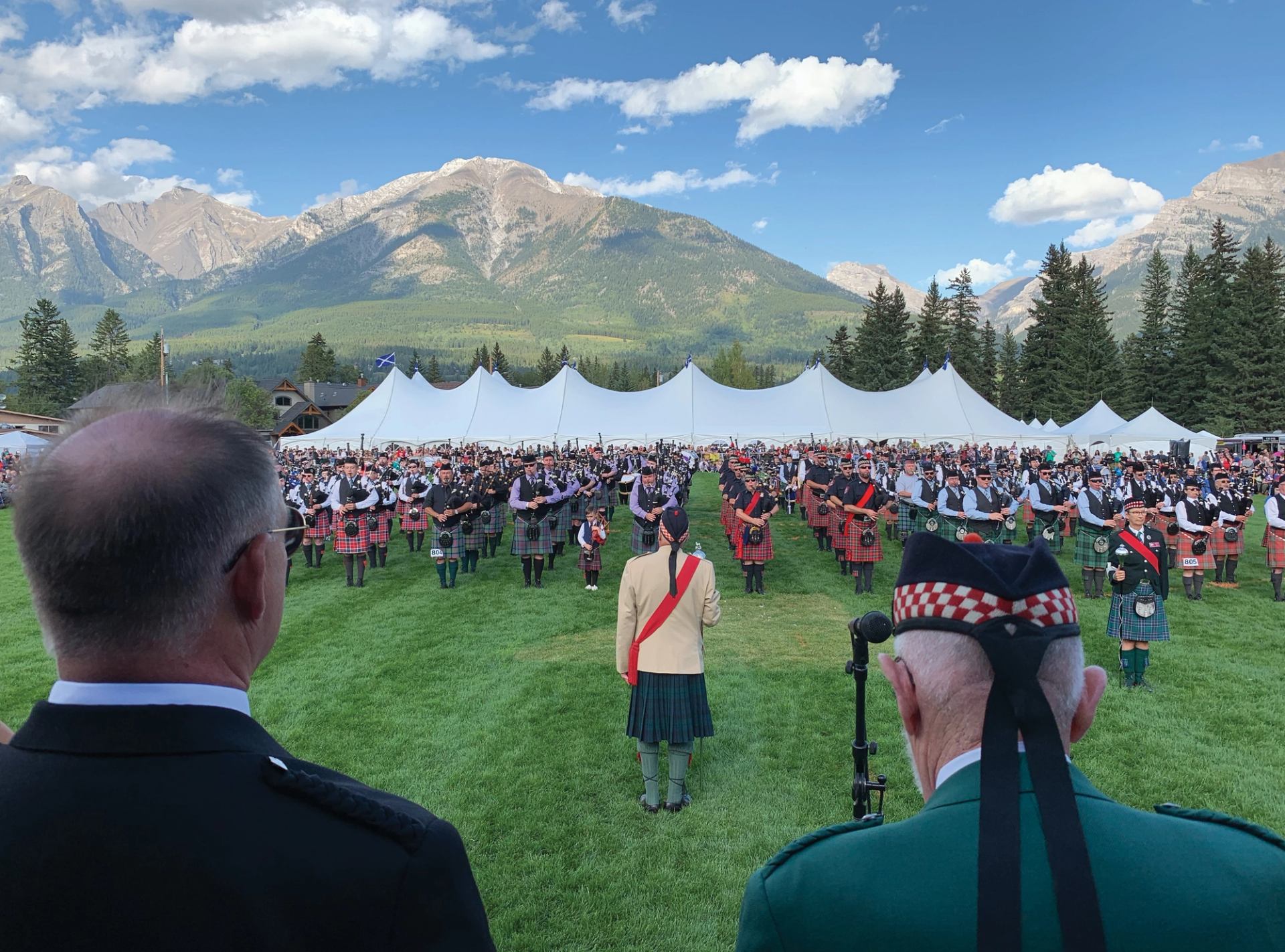 Massed pipe bands in tartan kilts perform on a field below Rocky Mountain peaks.