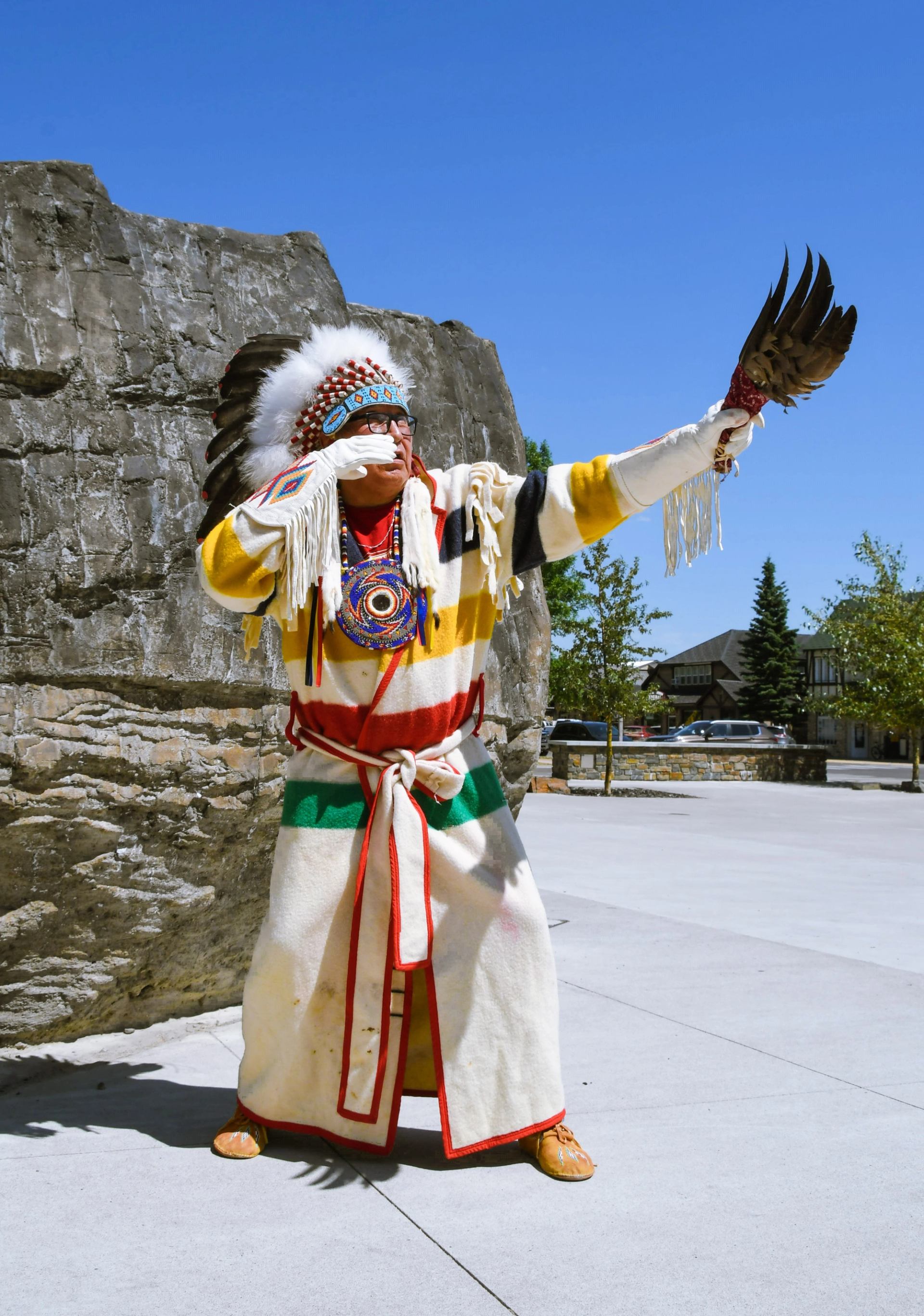 An Indigenous man in traditional regalia raises an eagle feather fan in a ceremonial gesture. He wears a headdress, beaded medallion, and a long robe with red, yellow, green, and black stripes, standing in front of a large rock on a sunny day.