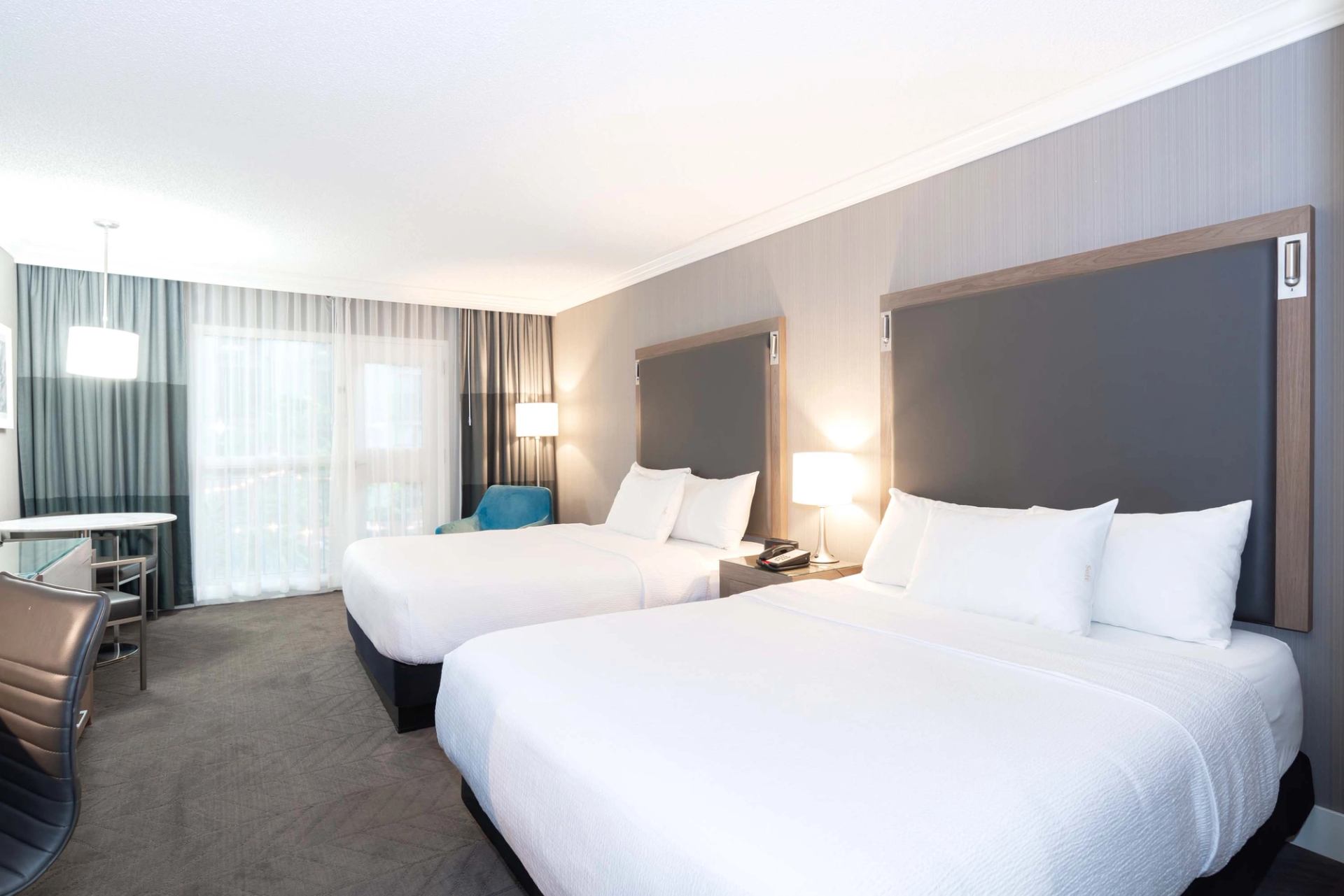 Hotel room with two double beds, grey headboards, desk, chair and window curtains.