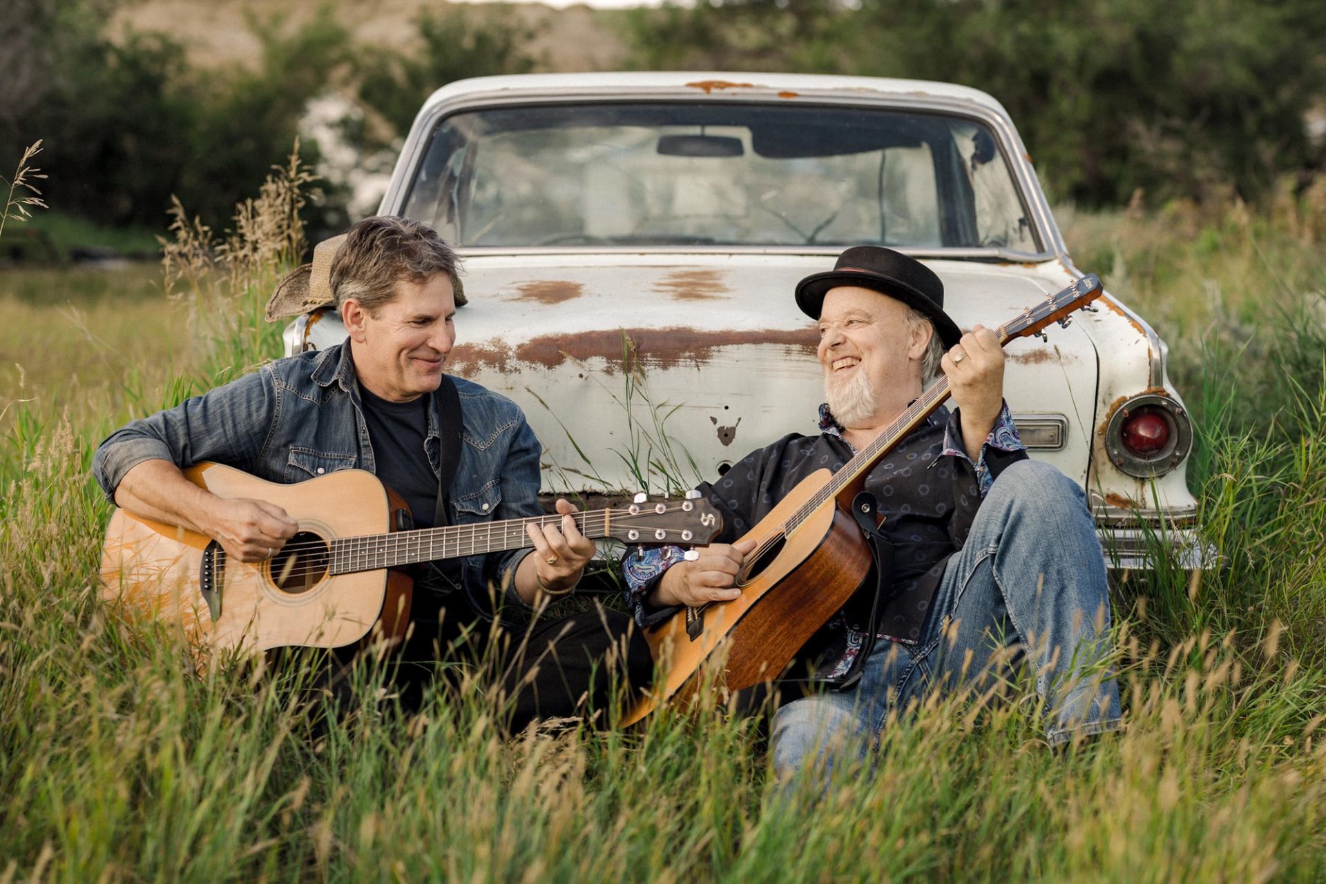 Two people sit in tall grass, playing acoustic guitars beside a rusty car.