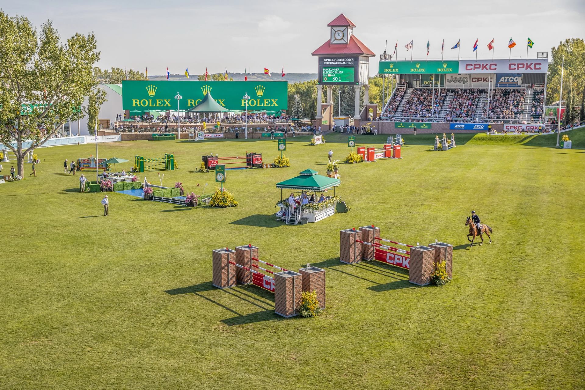 Wide view of the Spruce Meadows ‘Masters’ arena with jumps, spectators, and a rider on course.