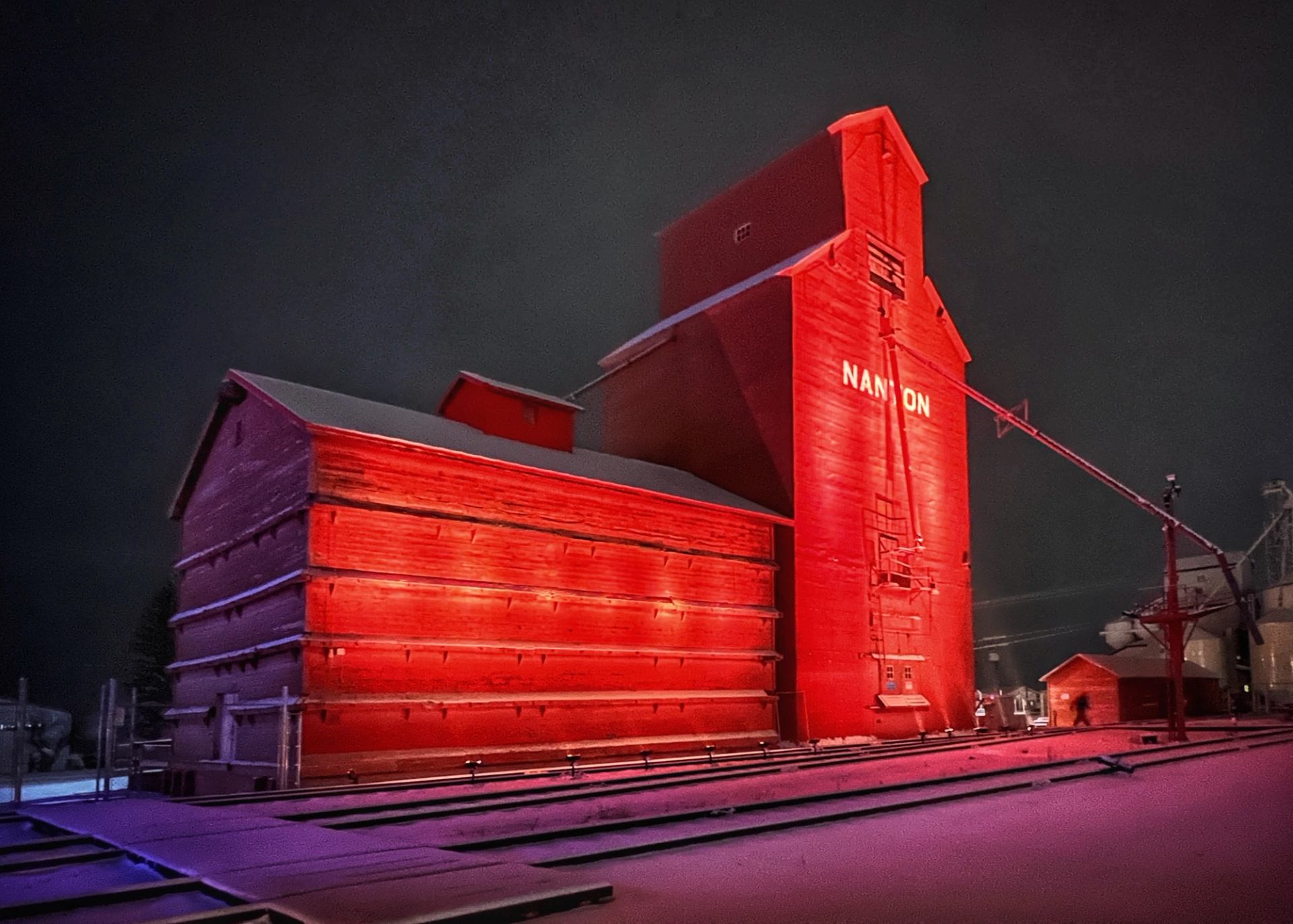 Red-lit grain elevator standing beside snowy tracks at night.