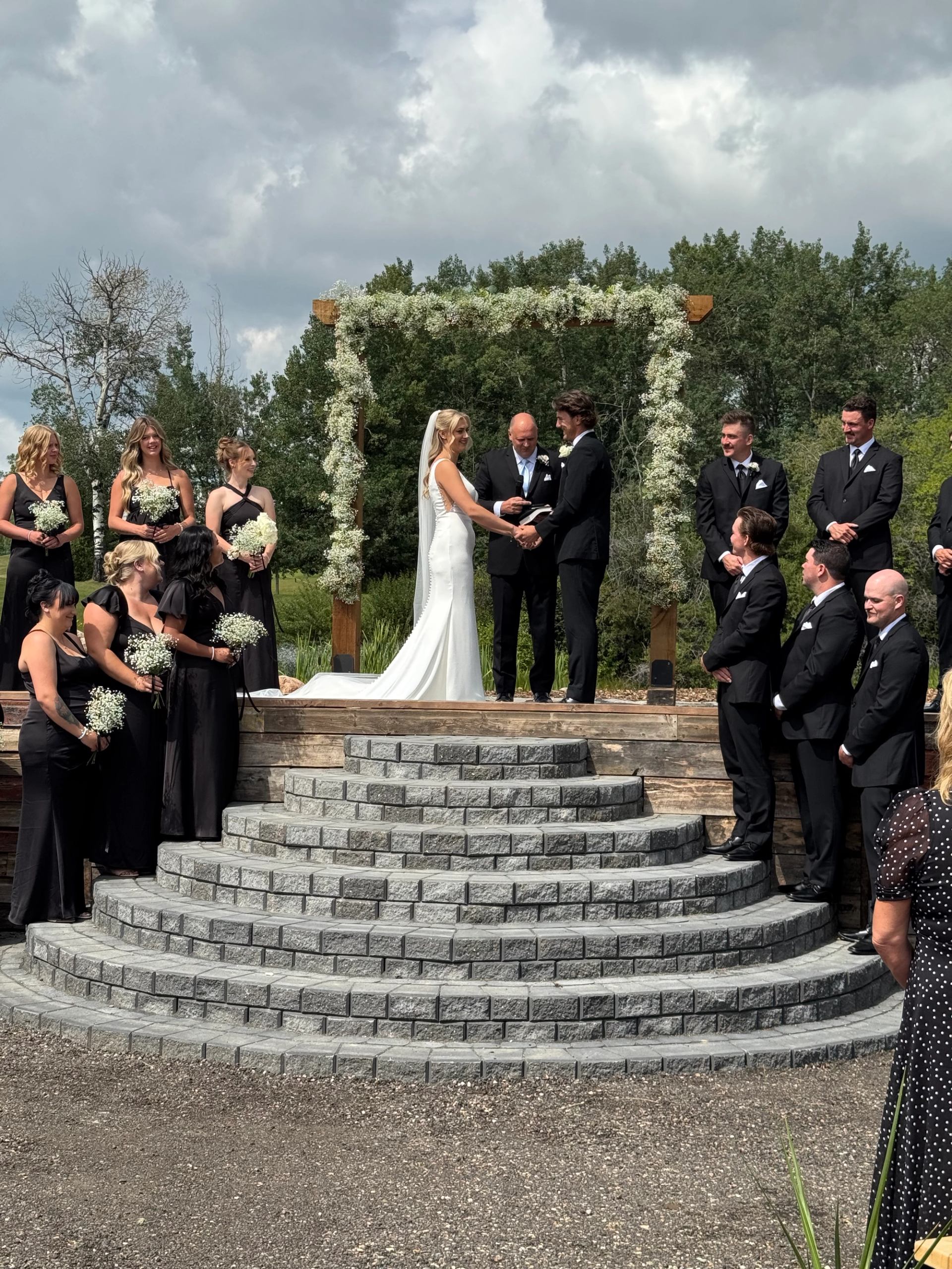 Outdoor wedding ceremony on stone platform under greenery-covered arch.