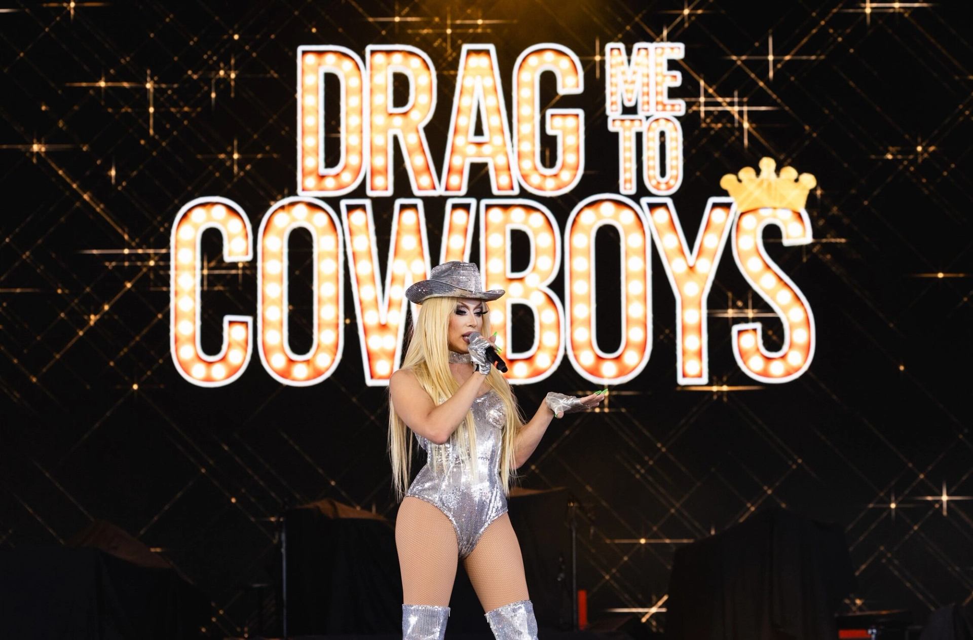 Drag performer in silver outfit sings onstage before large Drag Me to Cowboys sign.