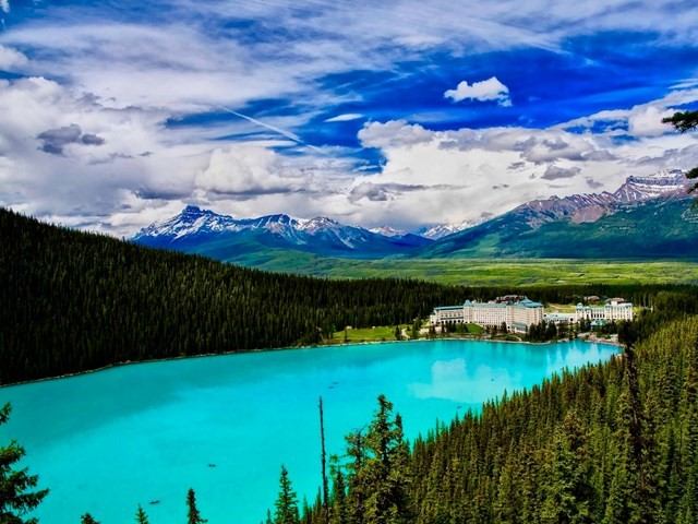 Turquoise lake surrounded by dense pine forest, with a large hotel on the shore and snow-capped mountains in the background under a partly cloudy sky.