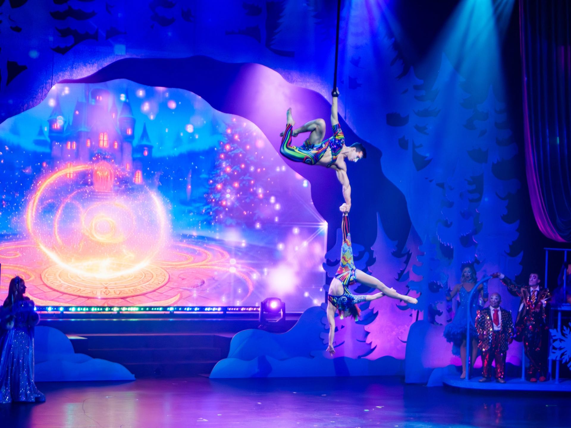 Aerial acrobats perform suspended above a stage with colourful lights and a fantasy backdrop.