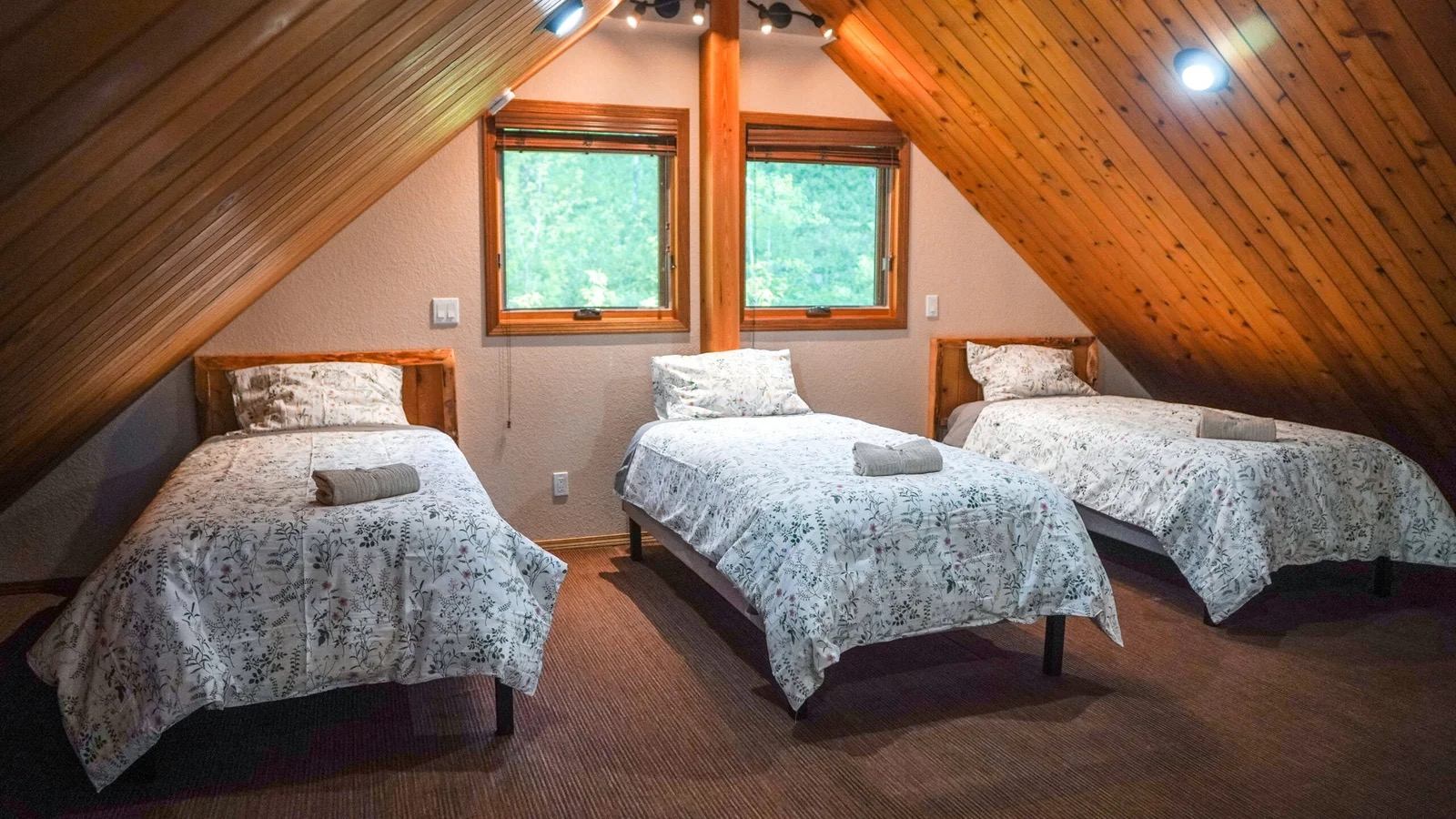 Wood-paneled cabin bedroom with three single beds, patterned bedding, and two windows.