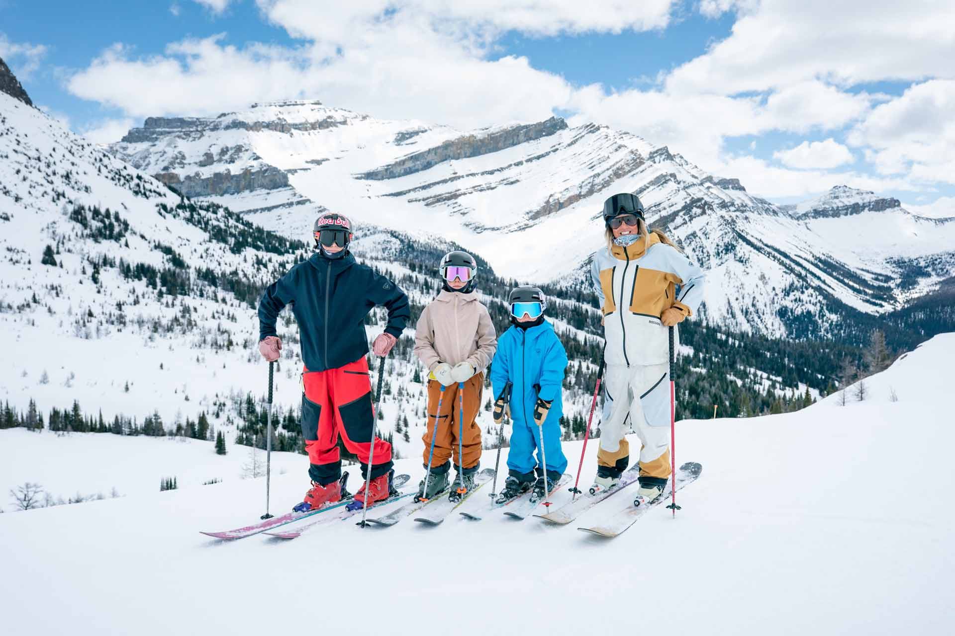 Skiers in colourful gear stand on snowy slope with mountain range and trees behind.