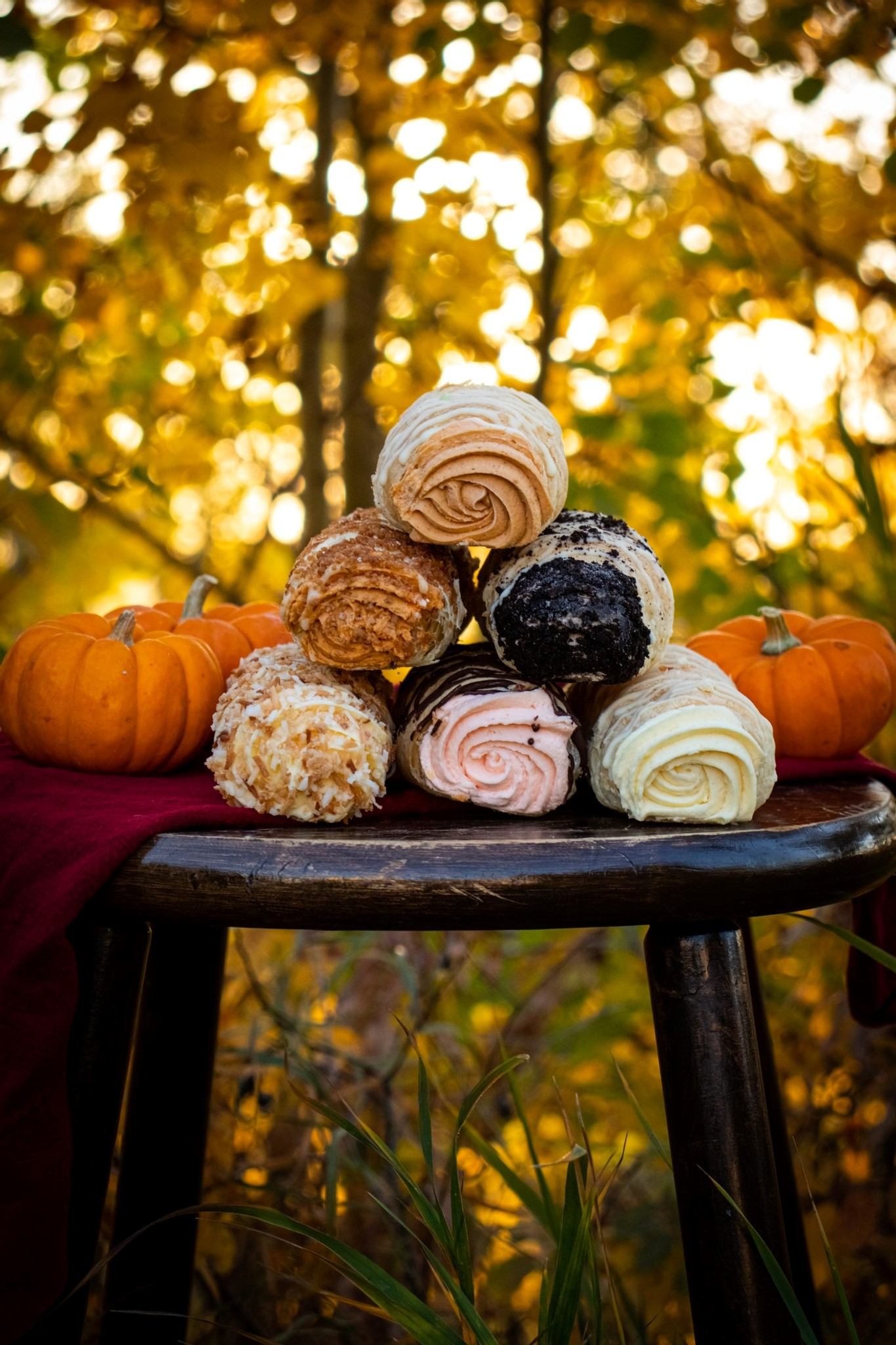 Stacked rolled pastries on wooden stool beside pumpkins, with blurred yellow leaves.