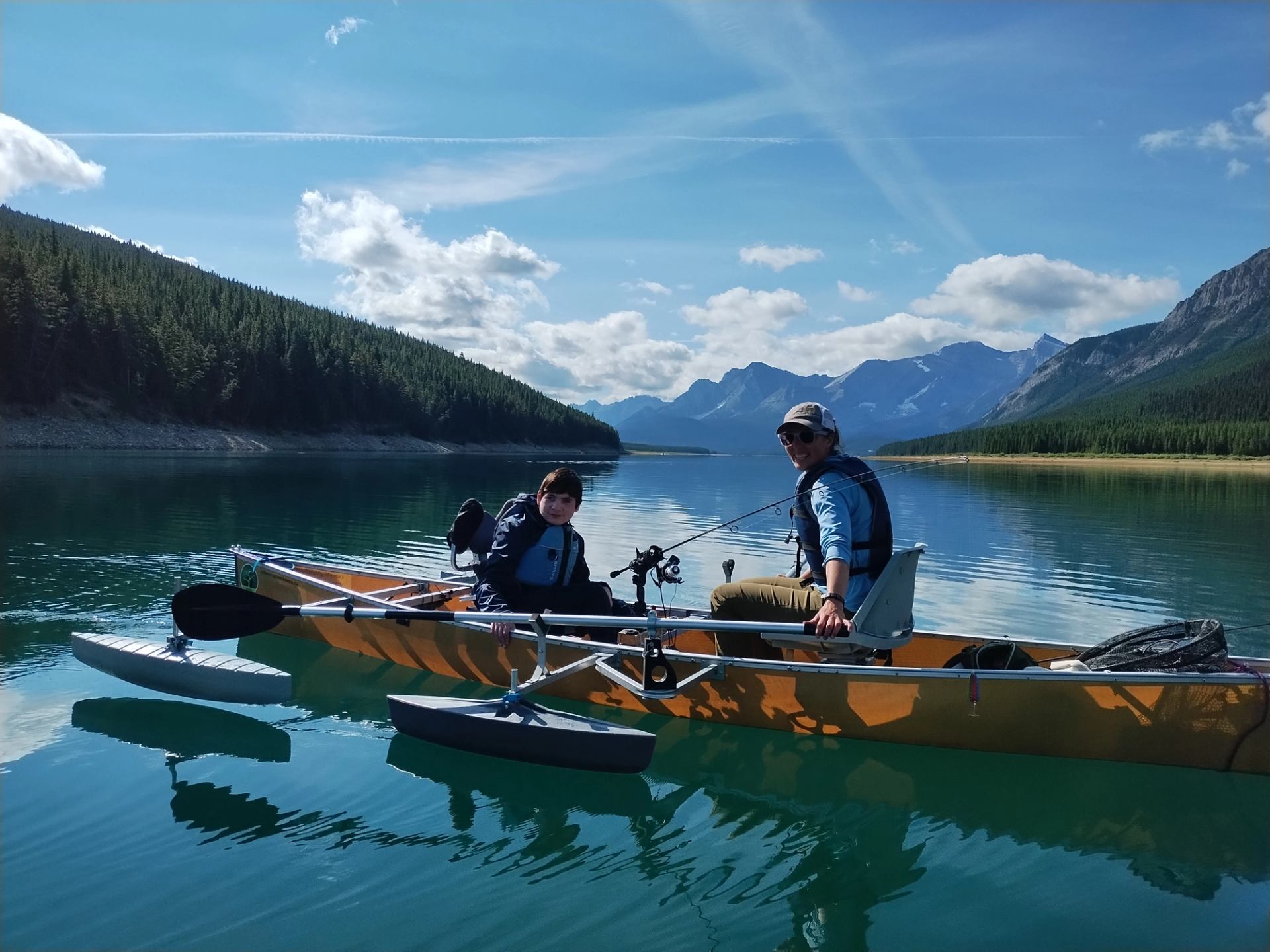 Two people in an adaptive kayak on a clear lake with mountains and forest in the distance.